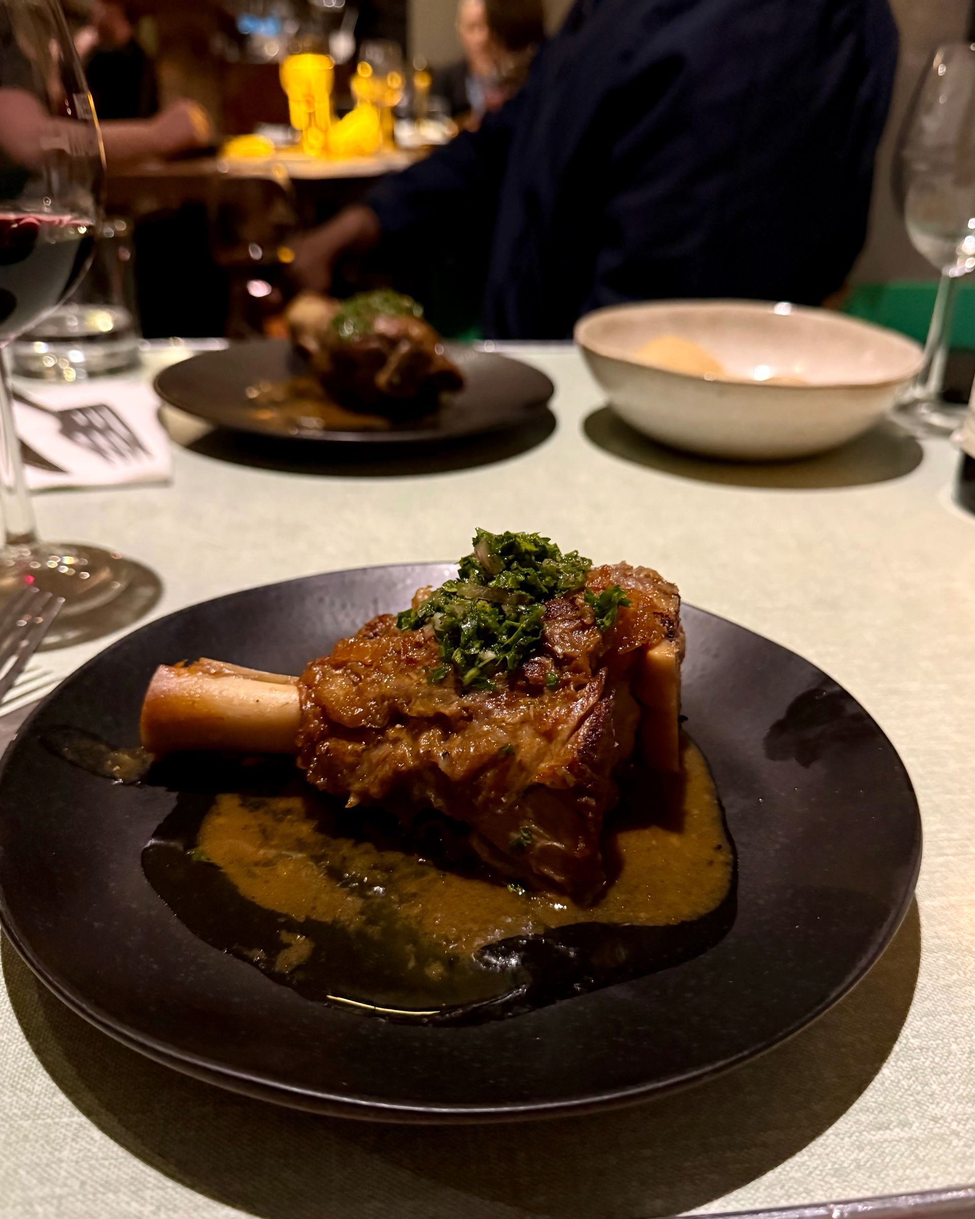 Braised meat shank on a black plate, drizzled with sauce and topped with herbs, on a restaurant table.