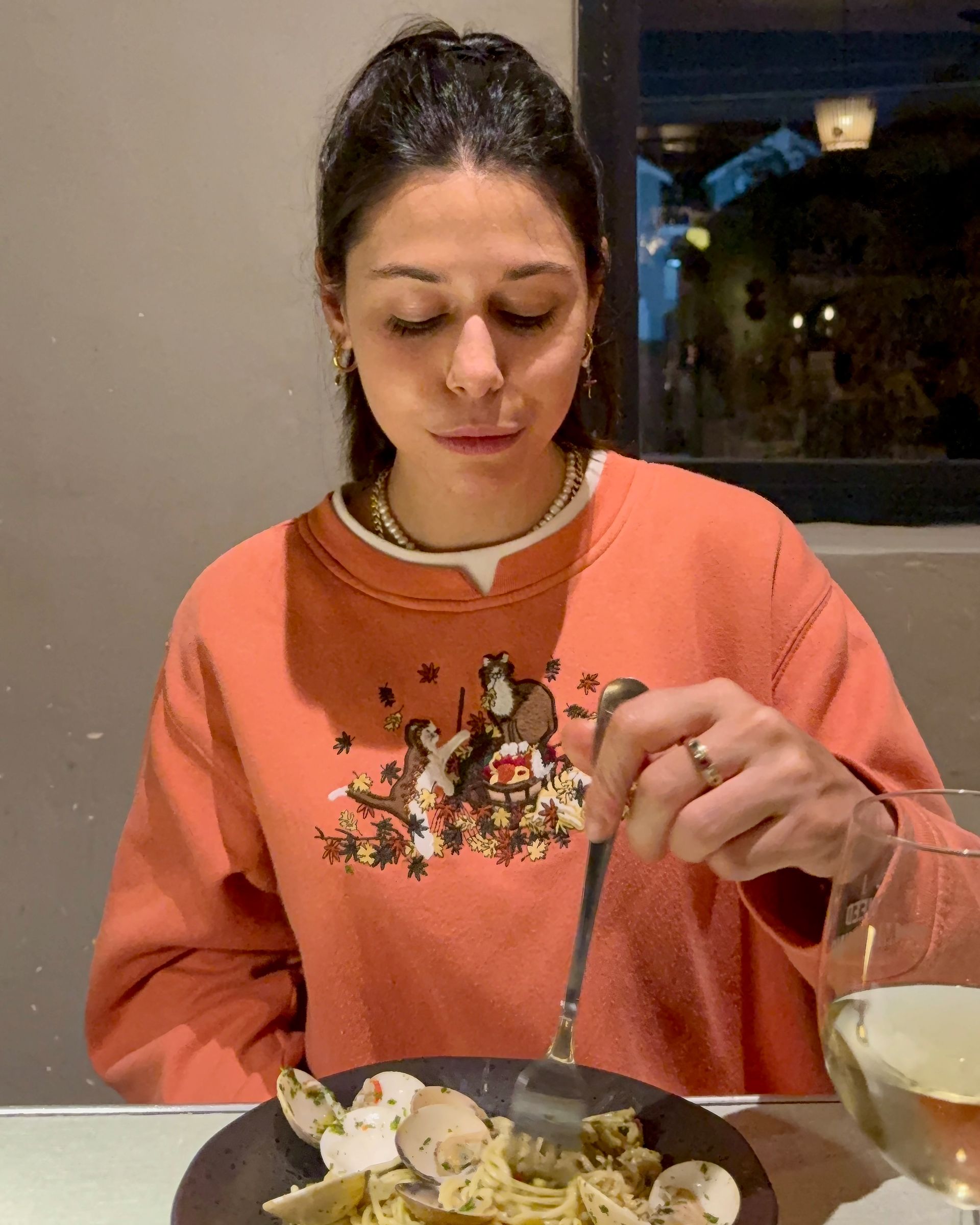 Woman in peach sweater eating pasta at a table, looking down, indoor setting.