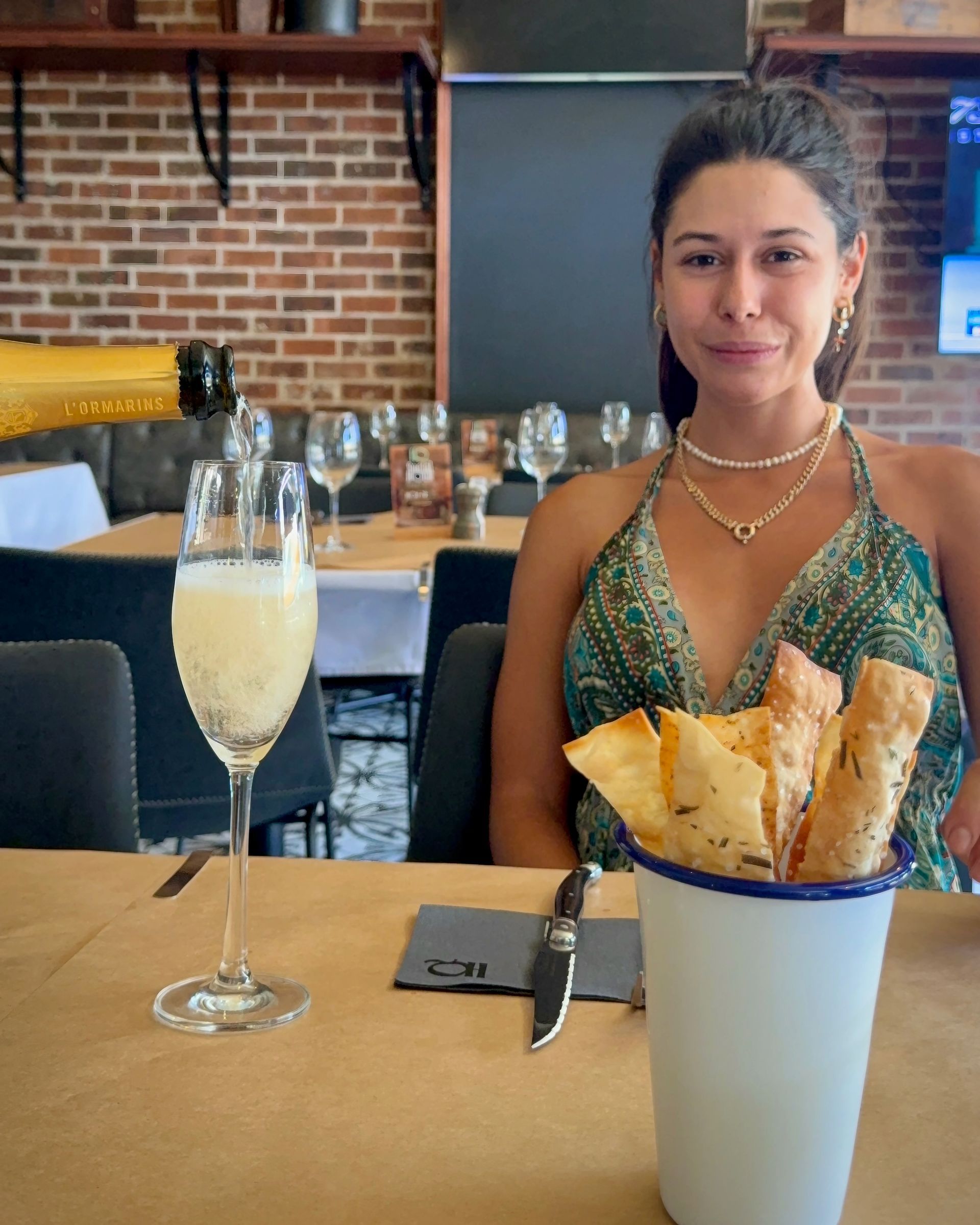 Woman at a restaurant table smiles, with breadsticks, champagne, and glasses visible.