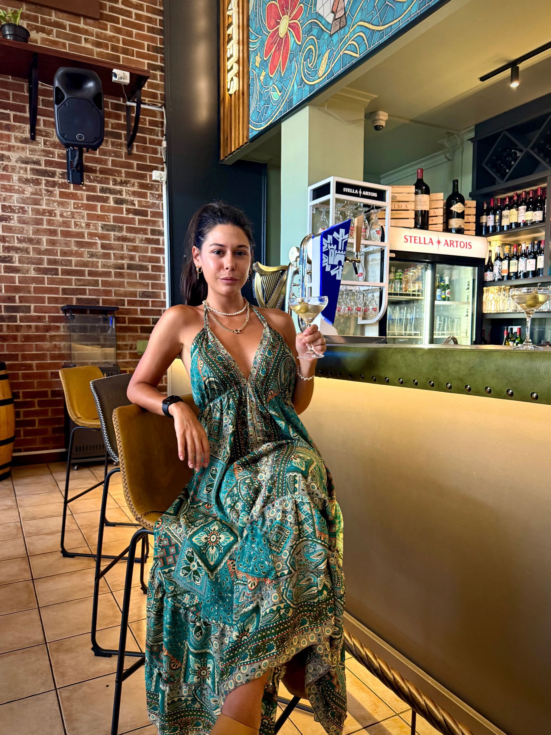 Woman in green dress, sitting at a bar, holding a drink. Brick wall and bar backdrop.