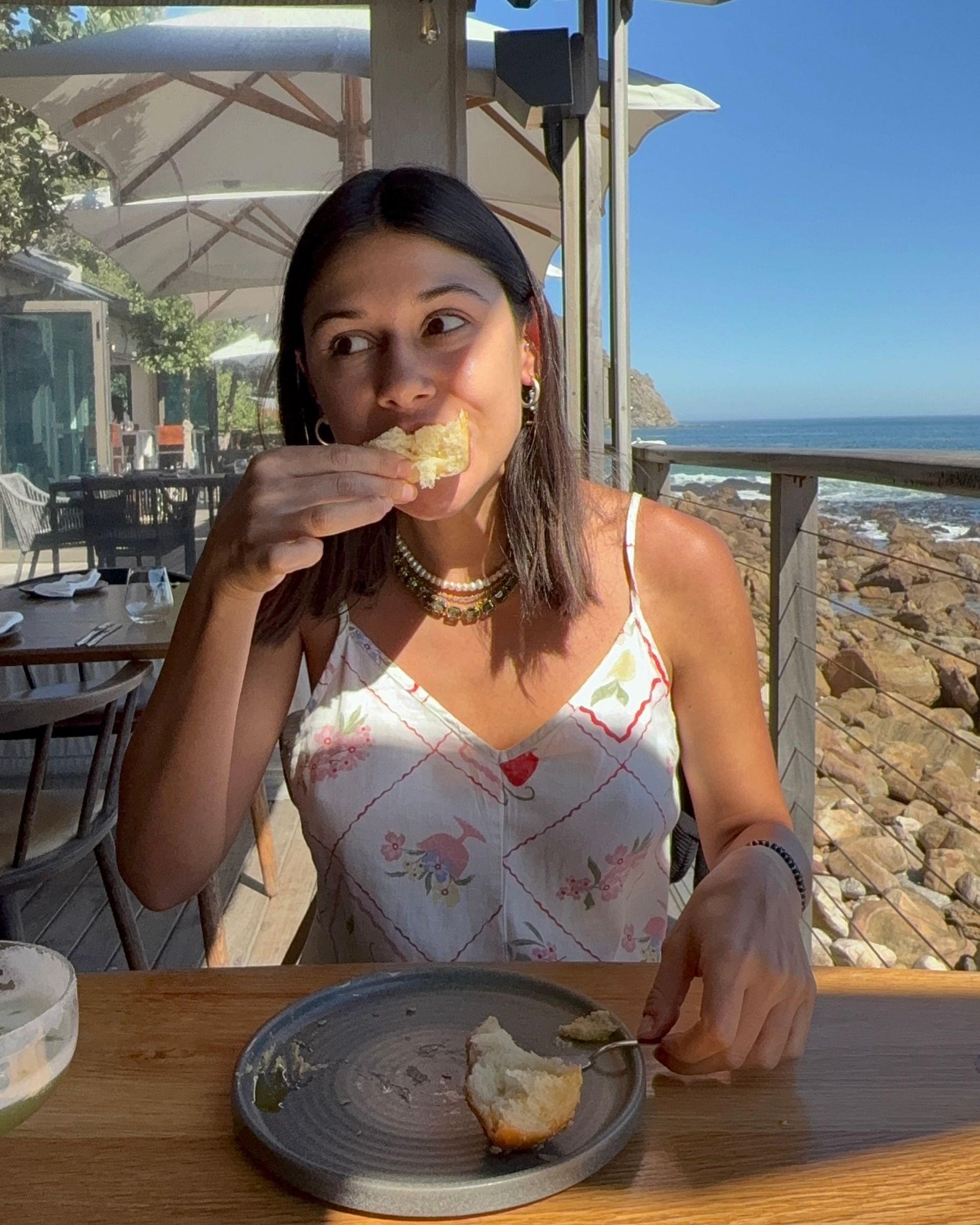 Woman eating bread outdoors at a restaurant near the ocean on a sunny day.
