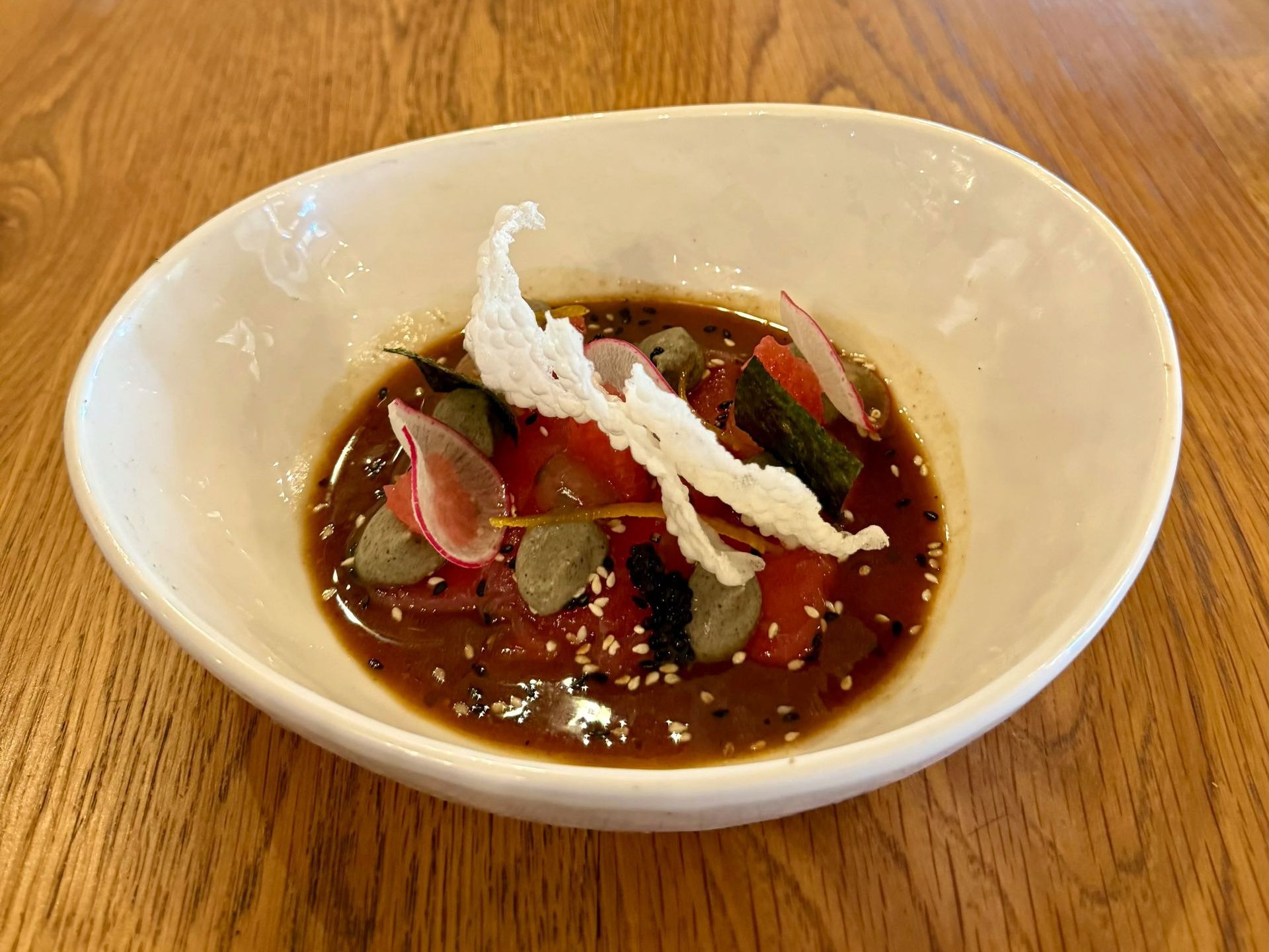 White bowl with a dark sauce, topped with tomatoes, radishes, and a crispy white garnish.