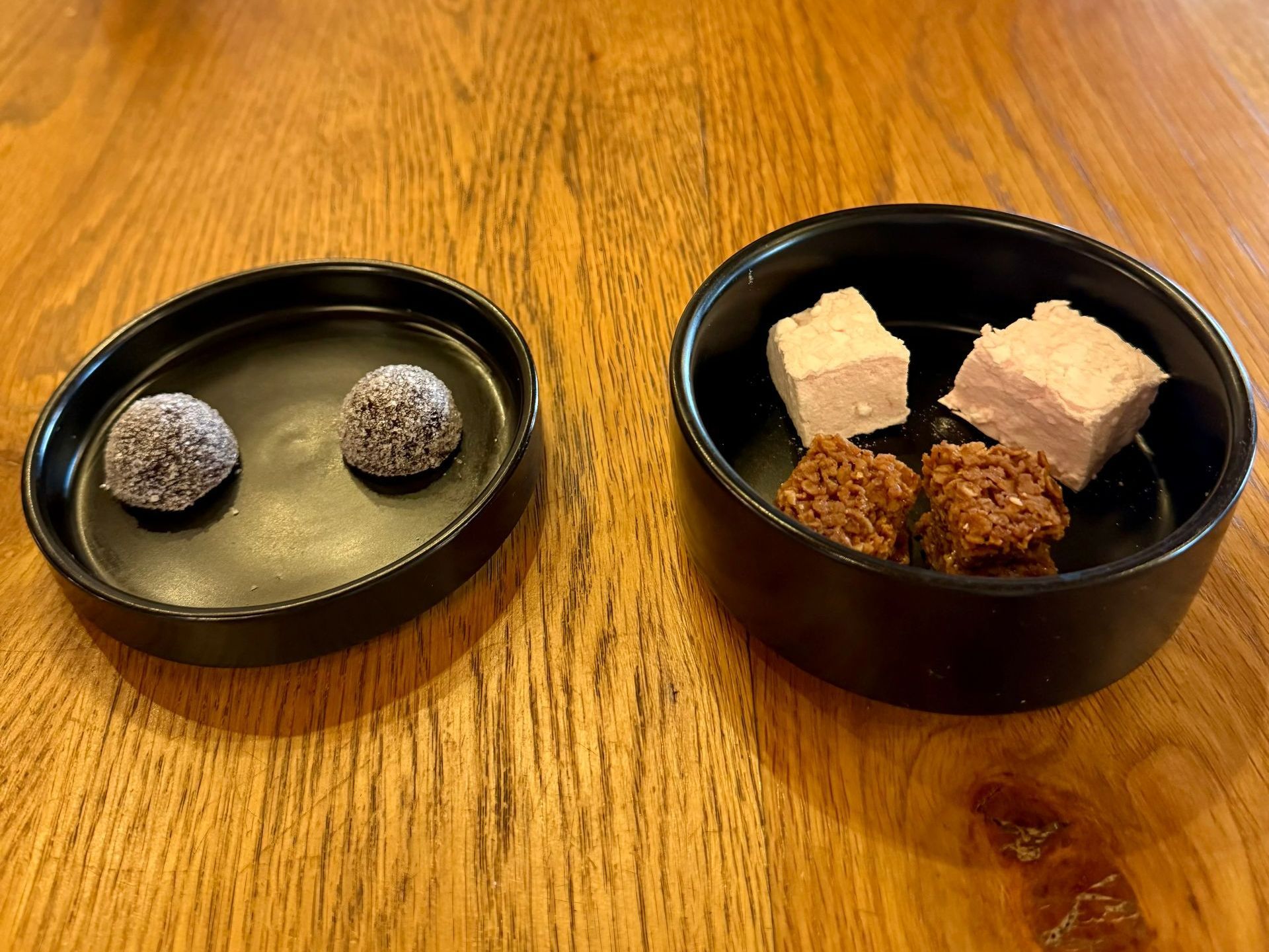 Two black bowls of desserts on a wooden table. One has two truffles, the other has marshmallows and brownie bites.