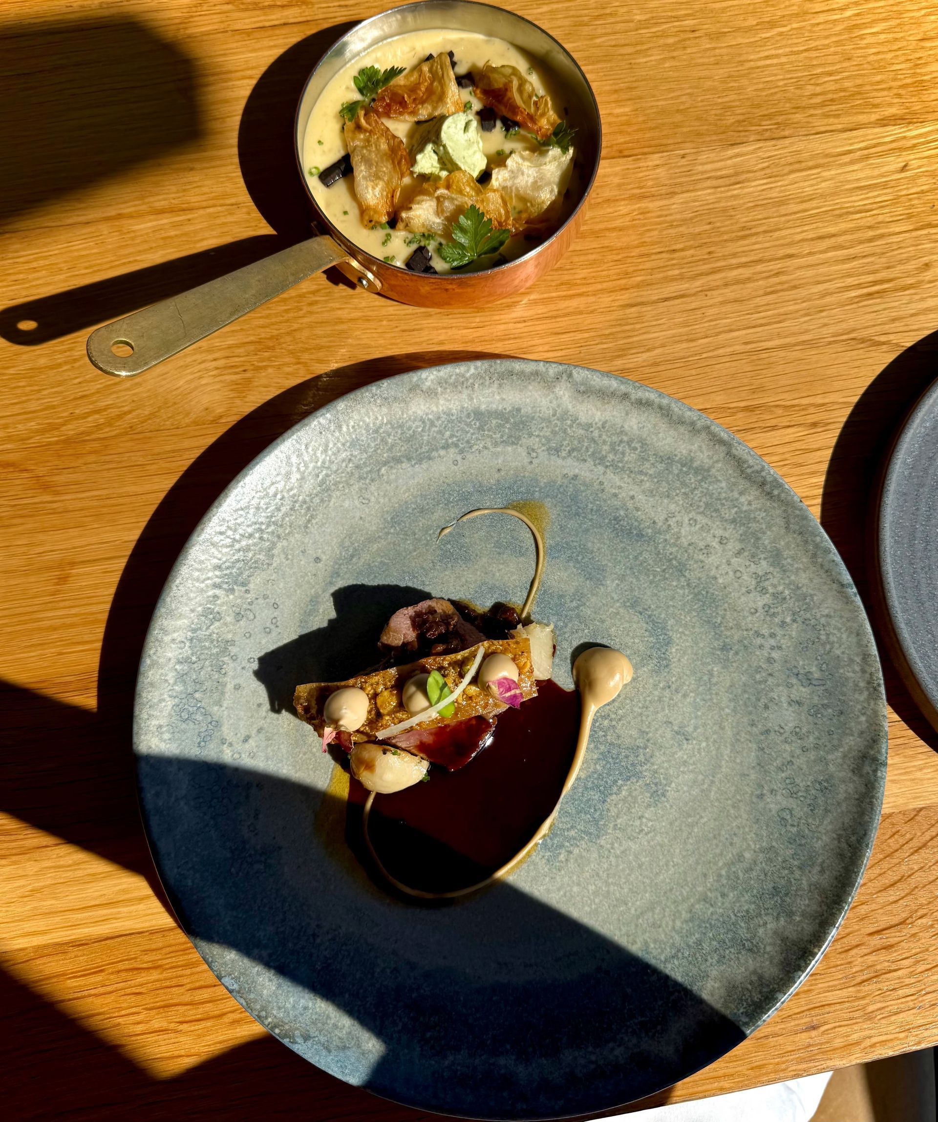 Gourmet dish on a blue plate with red sauce, and soup in a copper pan on a wooden table.