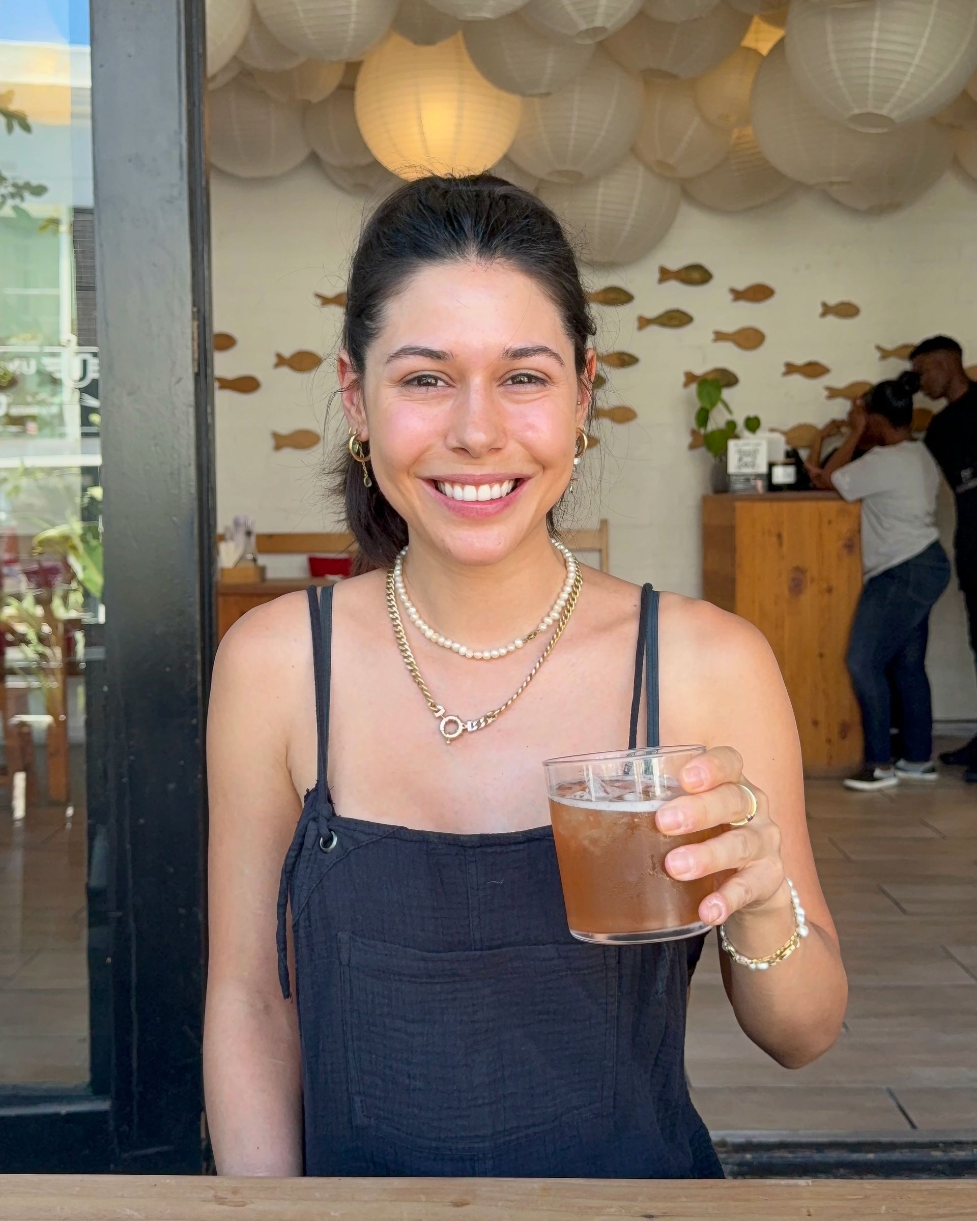 Woman smiling, holding a drink. Wearing a black dress, pearl necklaces, and gold jewelry. Outdoors, cafe setting.