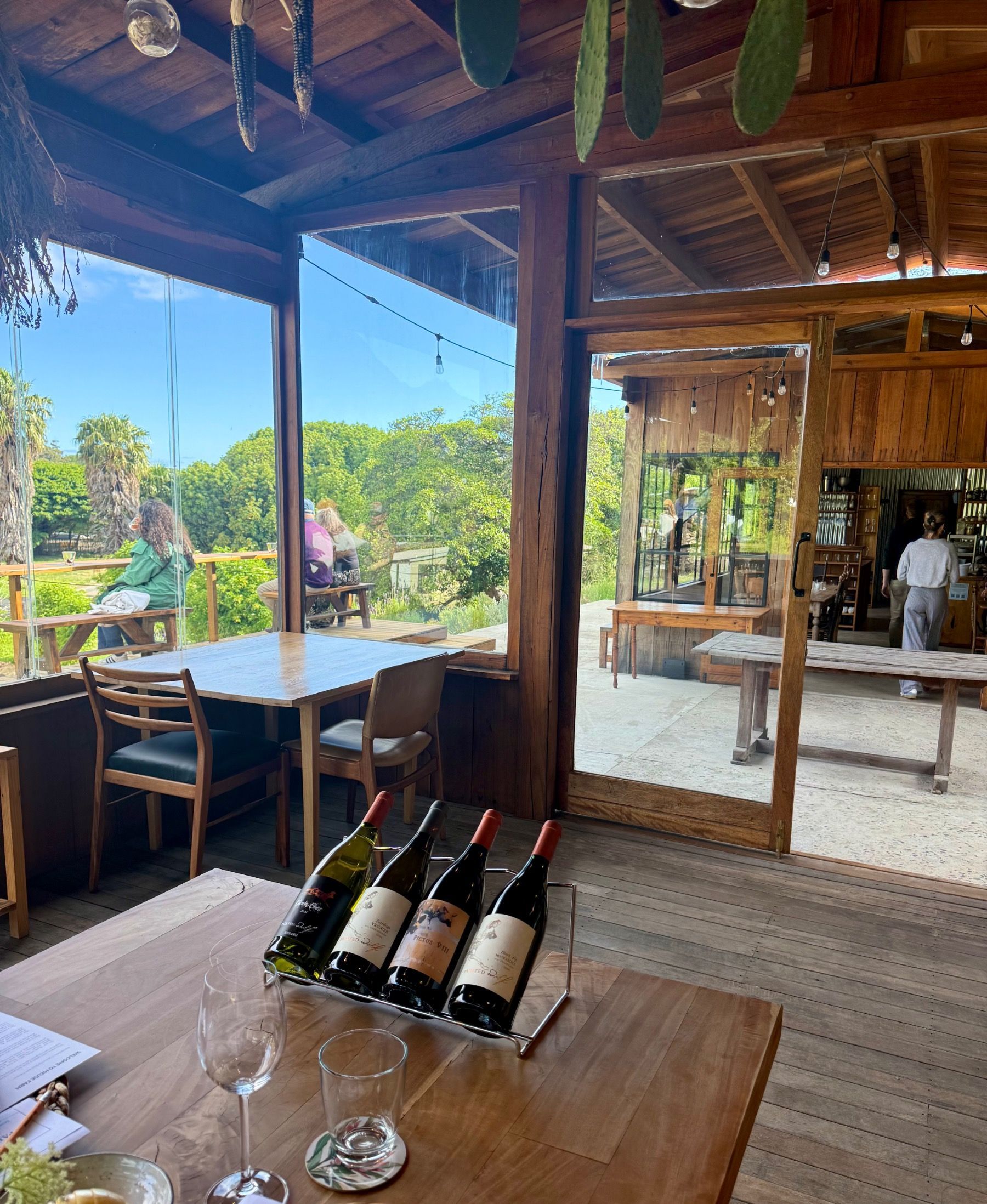 Long communal table with Painted Wolf Wines displayed at the head inside glass walled dining room at Meuse Farm