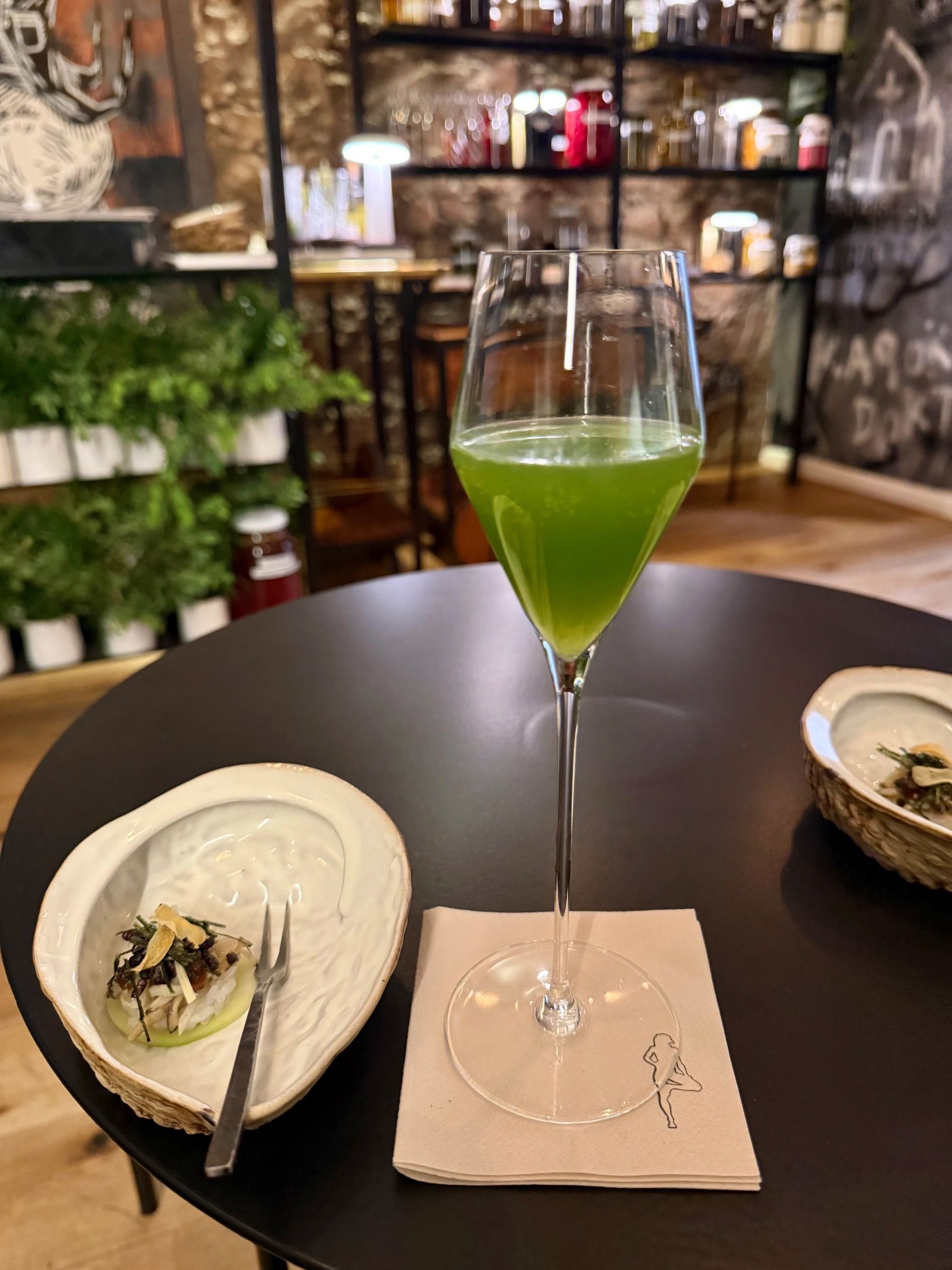 A green cocktail in a tall glass, next to a small appetizer on a black table.