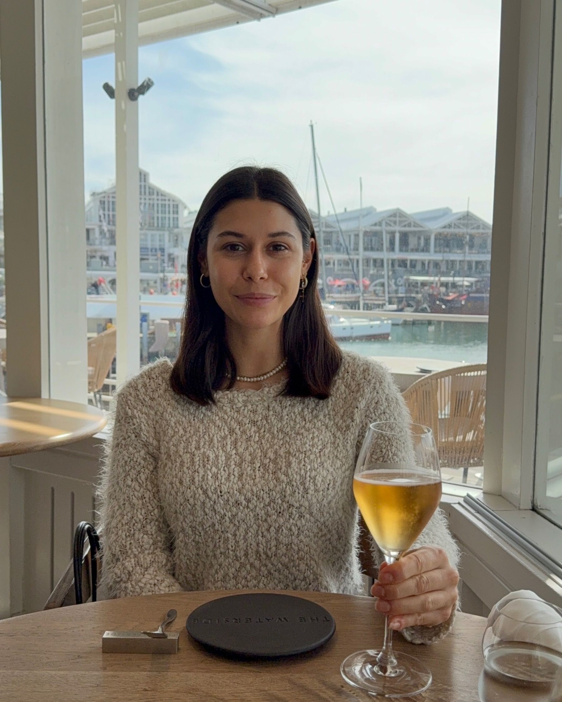 Paige holding a glass of rosé MCC inside The Waterside with harbour views behind her.
