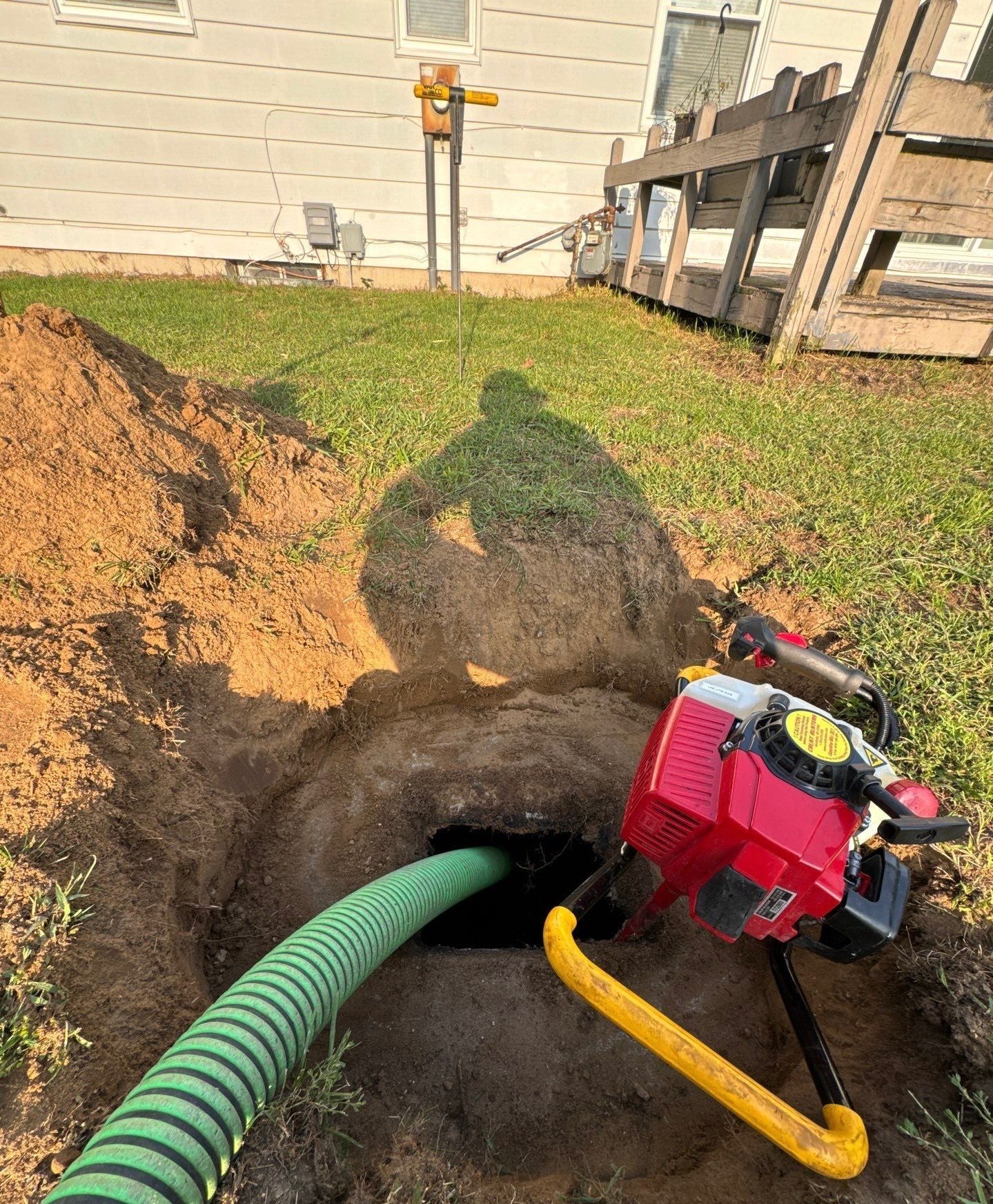 A hole in the ground with a green hose, red pump, and shadow cast on the grass.