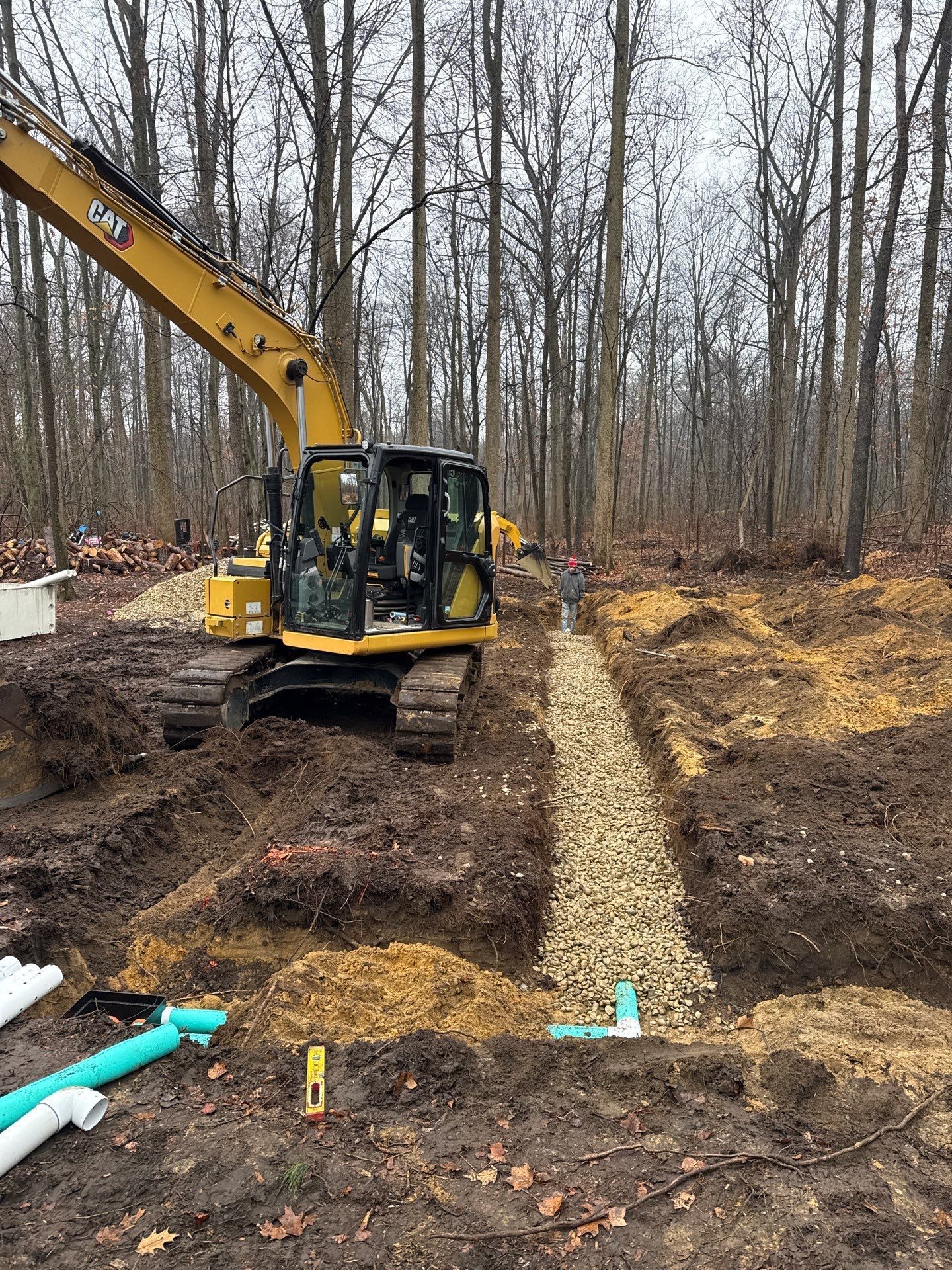 Yellow excavator digging a trench filled with gravel; pipes lie nearby in a wooded area.