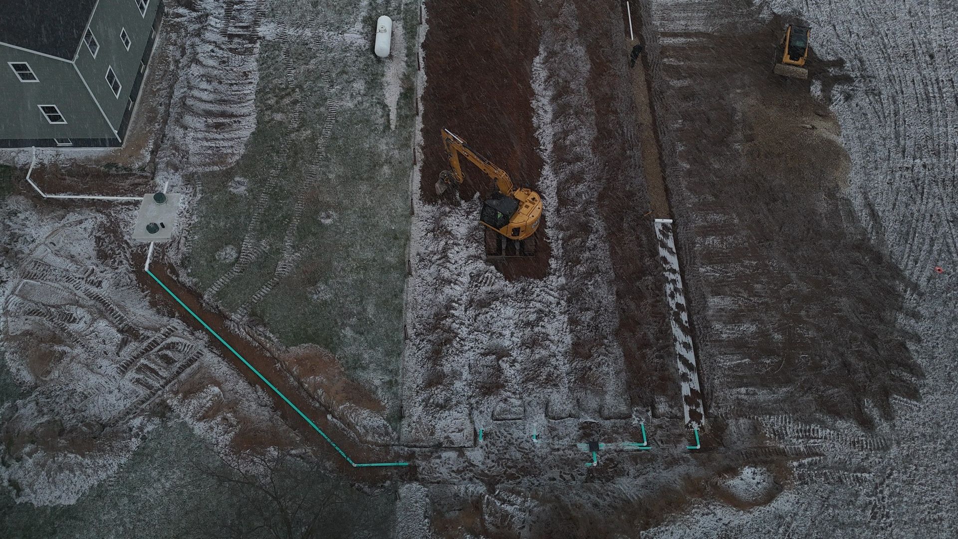 Overhead view of a construction site with an excavator in a muddy field next to a house, winter.
