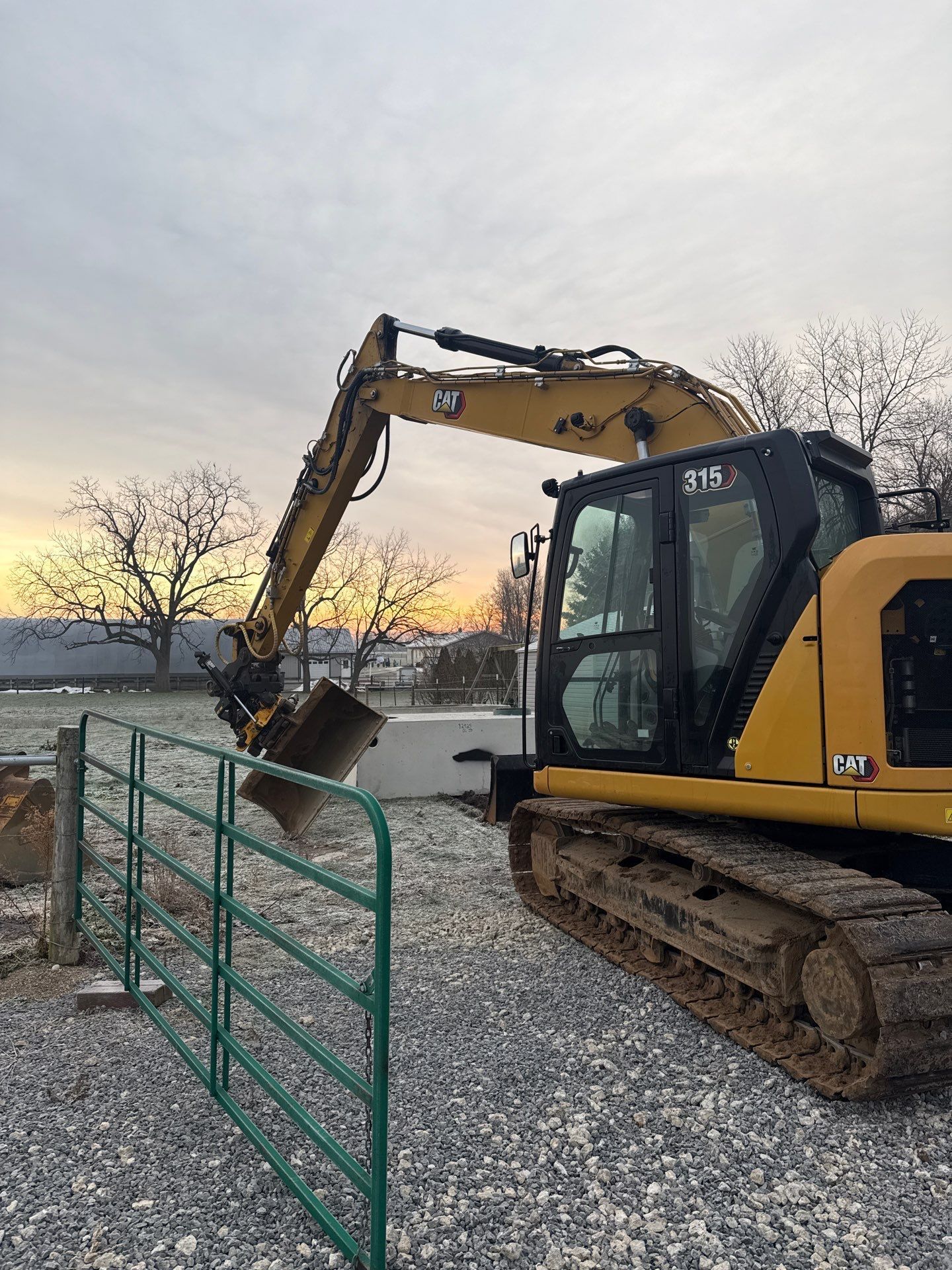 Yellow Caterpillar excavator digging near a green gate, against a cloudy sky.