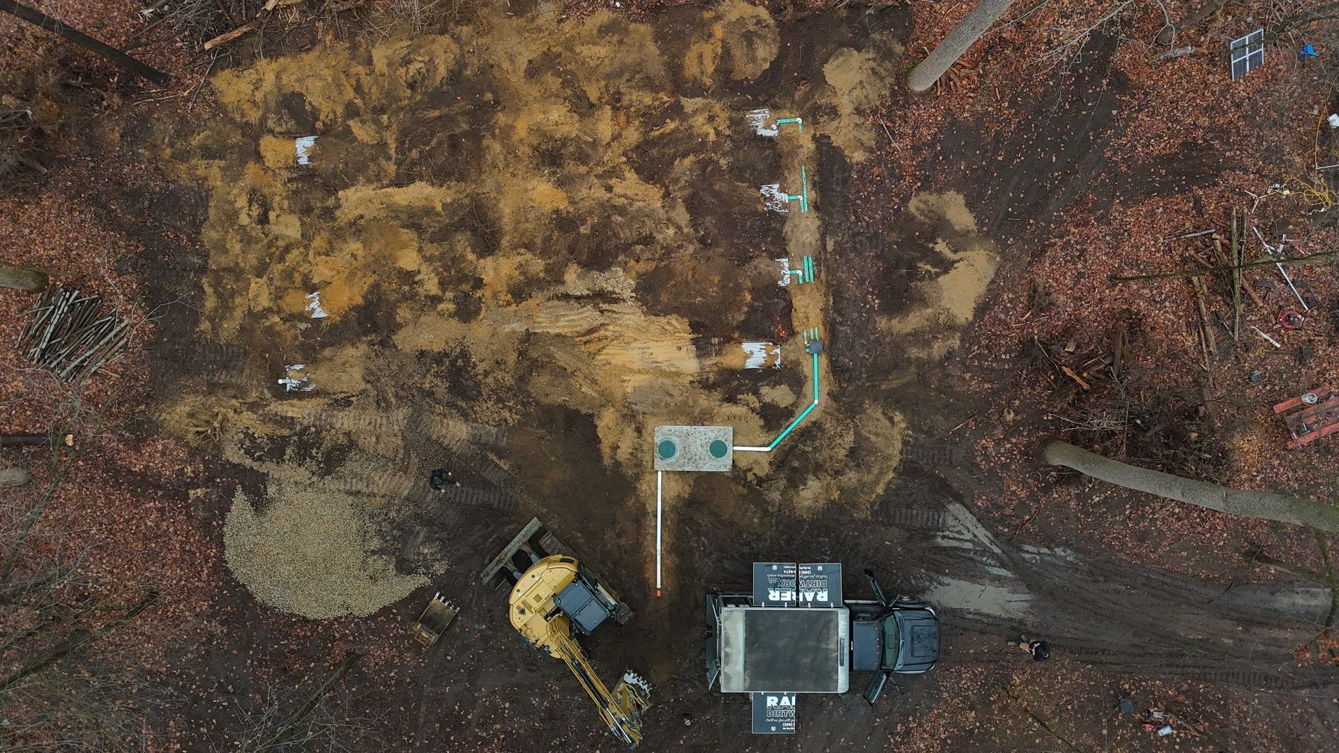 Overhead view of construction site in a forest. Excavator digging, dirt pile, equipment, and pipeline visible.