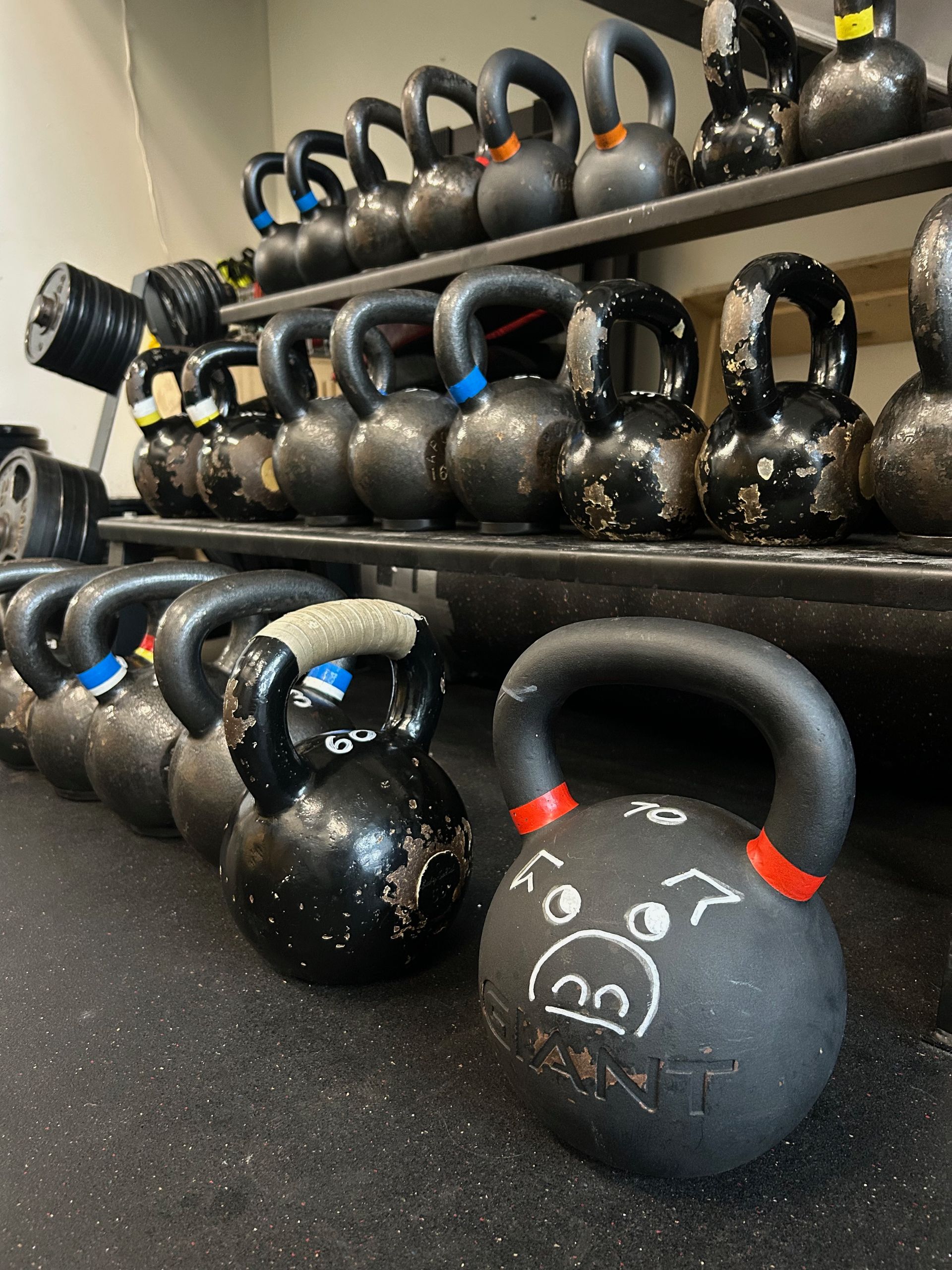 A rack and floor covered in black kettlebells, including one in the front decorated with a white chalk pig face drawing.