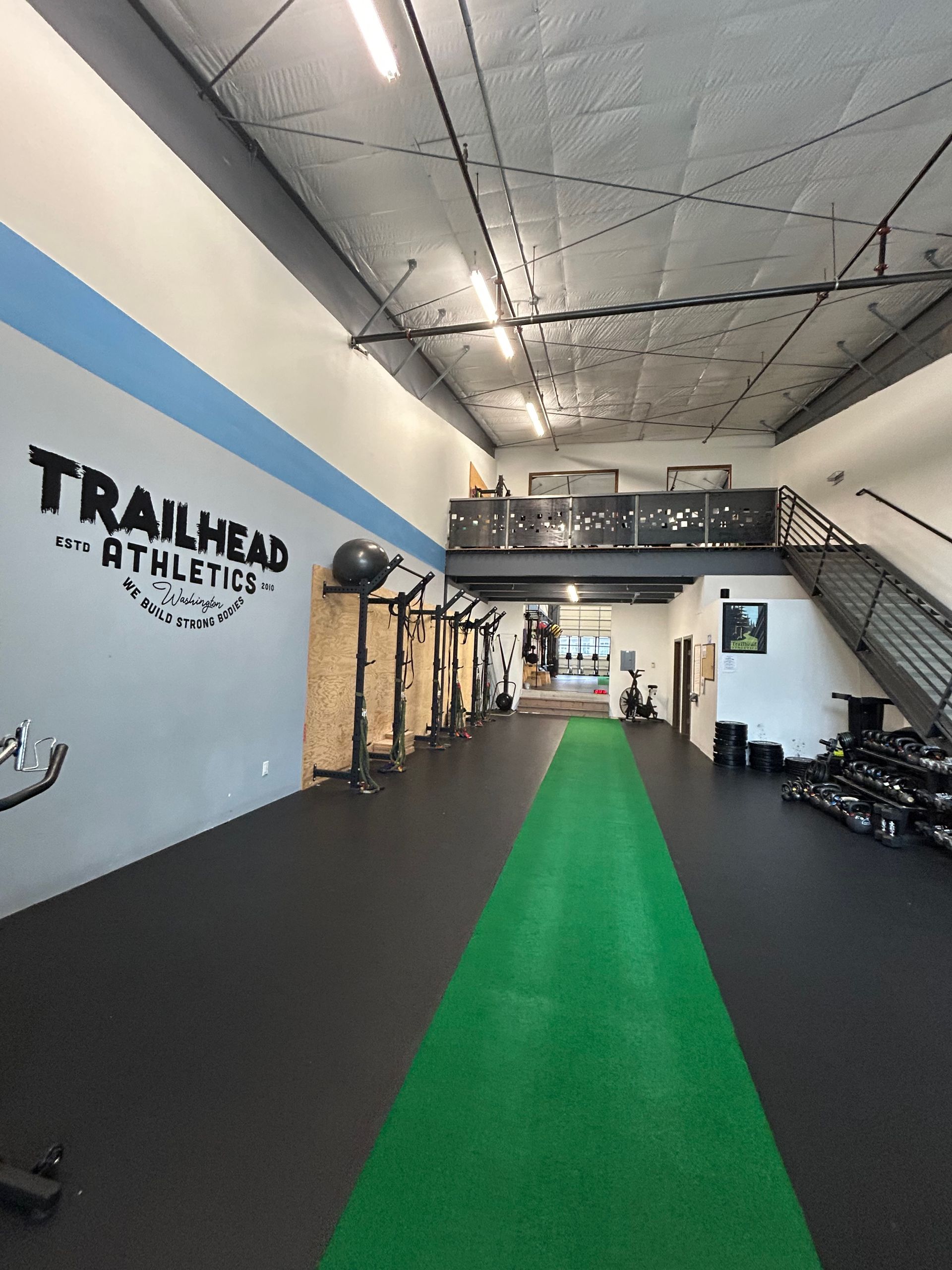 A gym interior with a central green turf track, black flooring, a mural logo on the left wall, and a staircase.