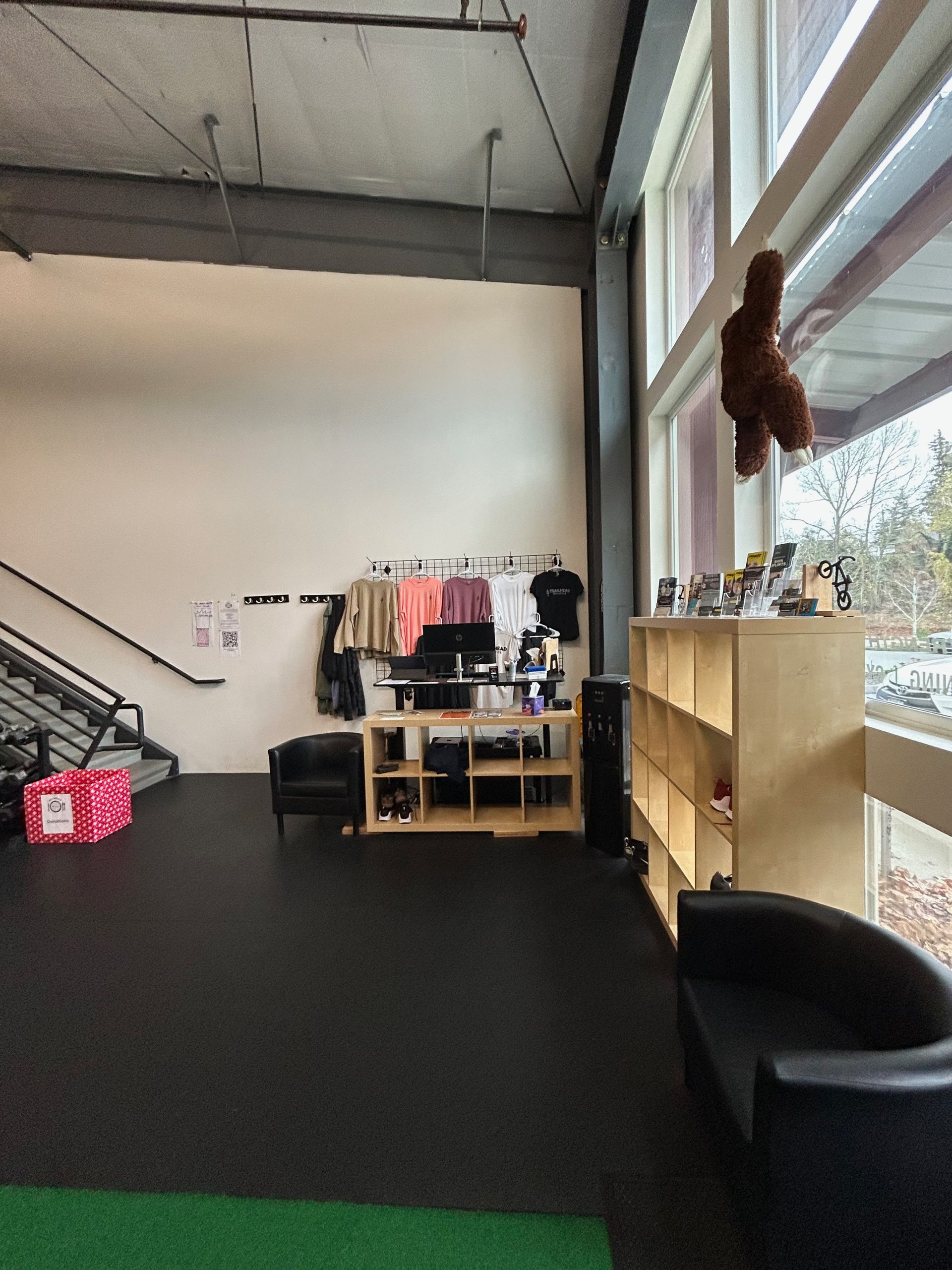 A bright gym lobby with a black floor, a wooden storage shelf, a hanging mascot, and a reception desk with apparel racks.