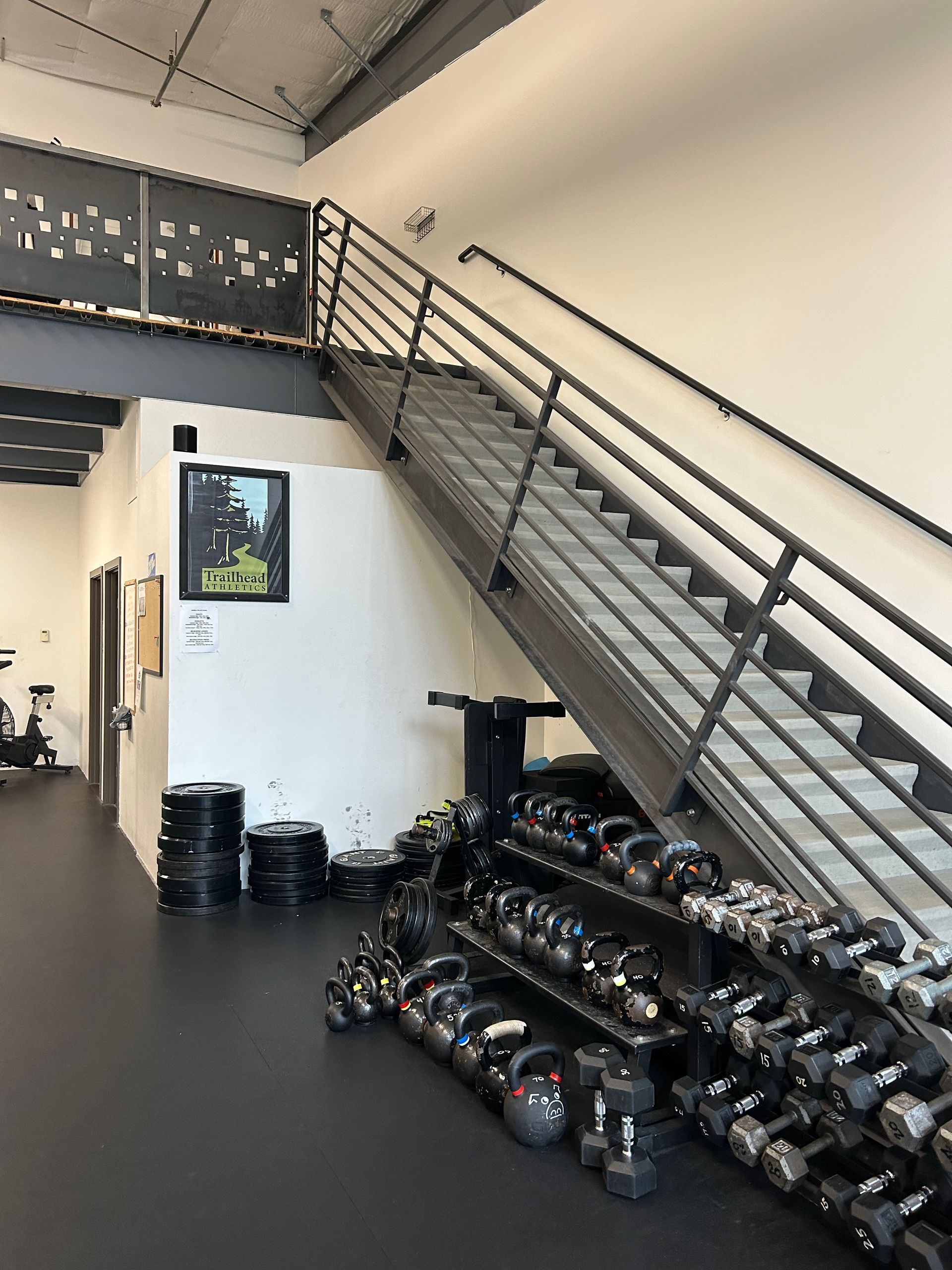 A metal staircase leads to a mezzanine in a gym, with a rack of various dumbbells and kettlebells stored underneath.