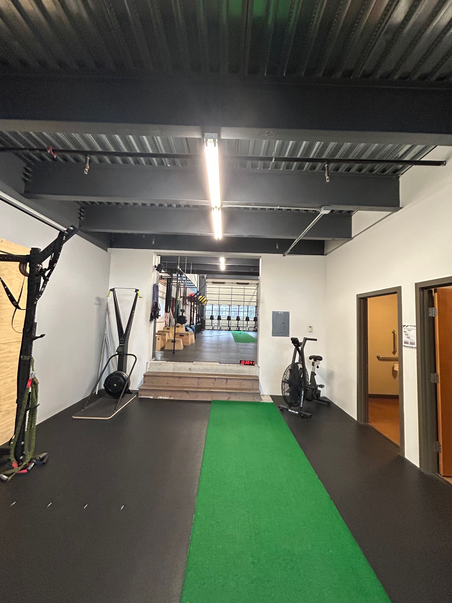 An indoor gym hallway with a central green turf runner, exercise equipment on the sides, and open doorways.