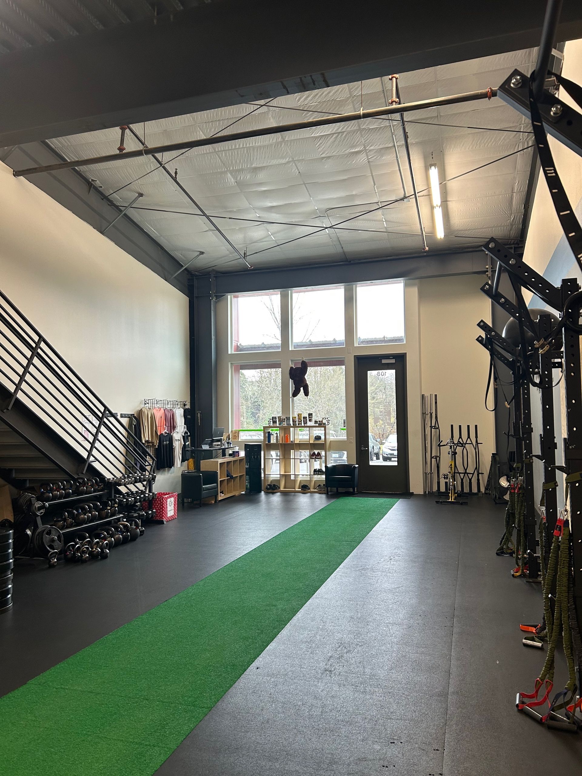 A high-ceilinged indoor fitness gym with a green turf strip in the center, gym equipment on the sides, and large windows.