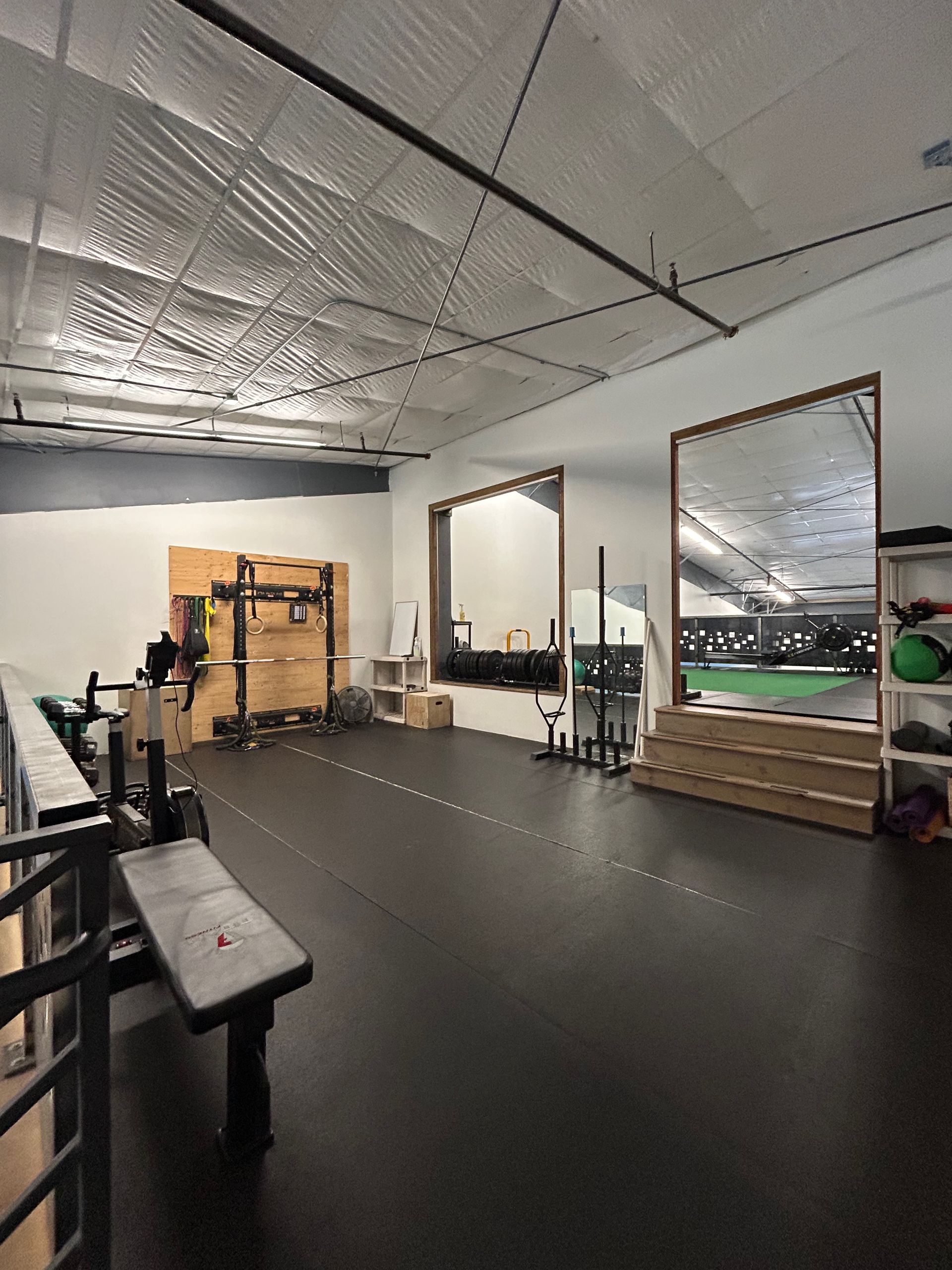 A modern home gym with a black floor, weight bench, squat rack, and large mirrors reflecting an indoor turf field.