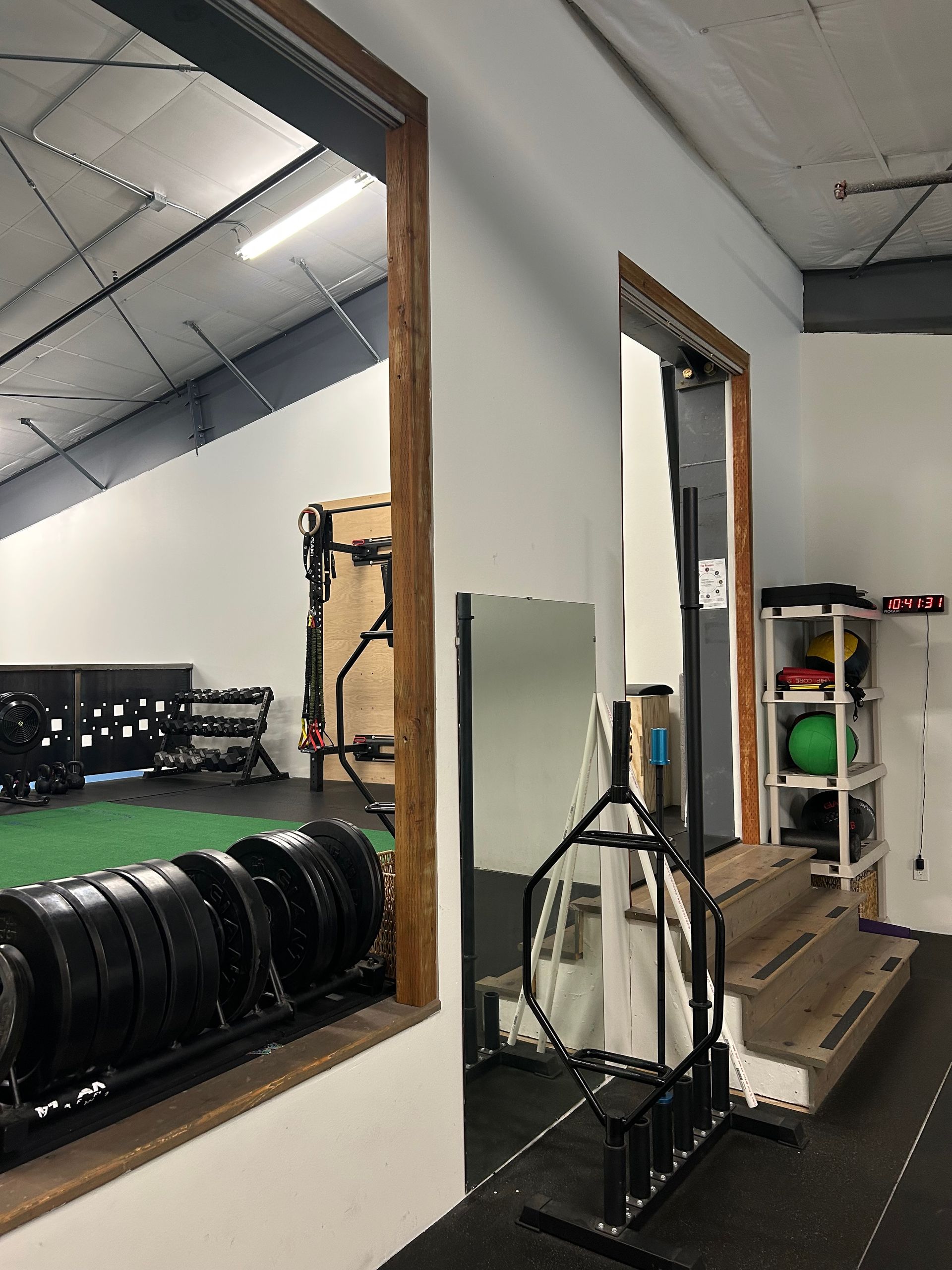 A gym interior featuring exercise equipment, heavy plates on racks, and stairs leading to a doorway reflected in a mirror.