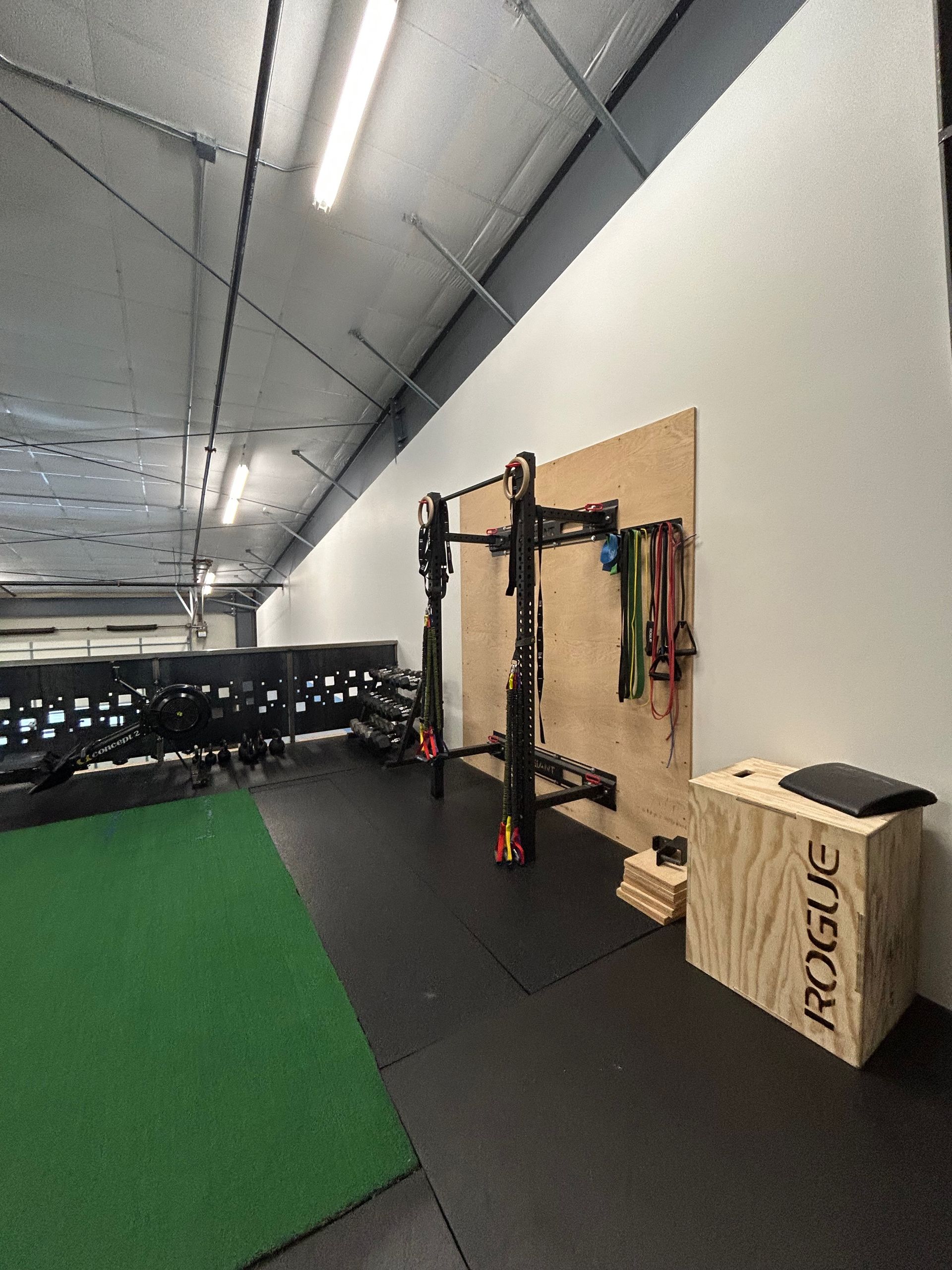 A gym space featuring a wall-mounted squat rack, colorful resistance bands, a Rogue wooden plyo box, and green turf flooring.