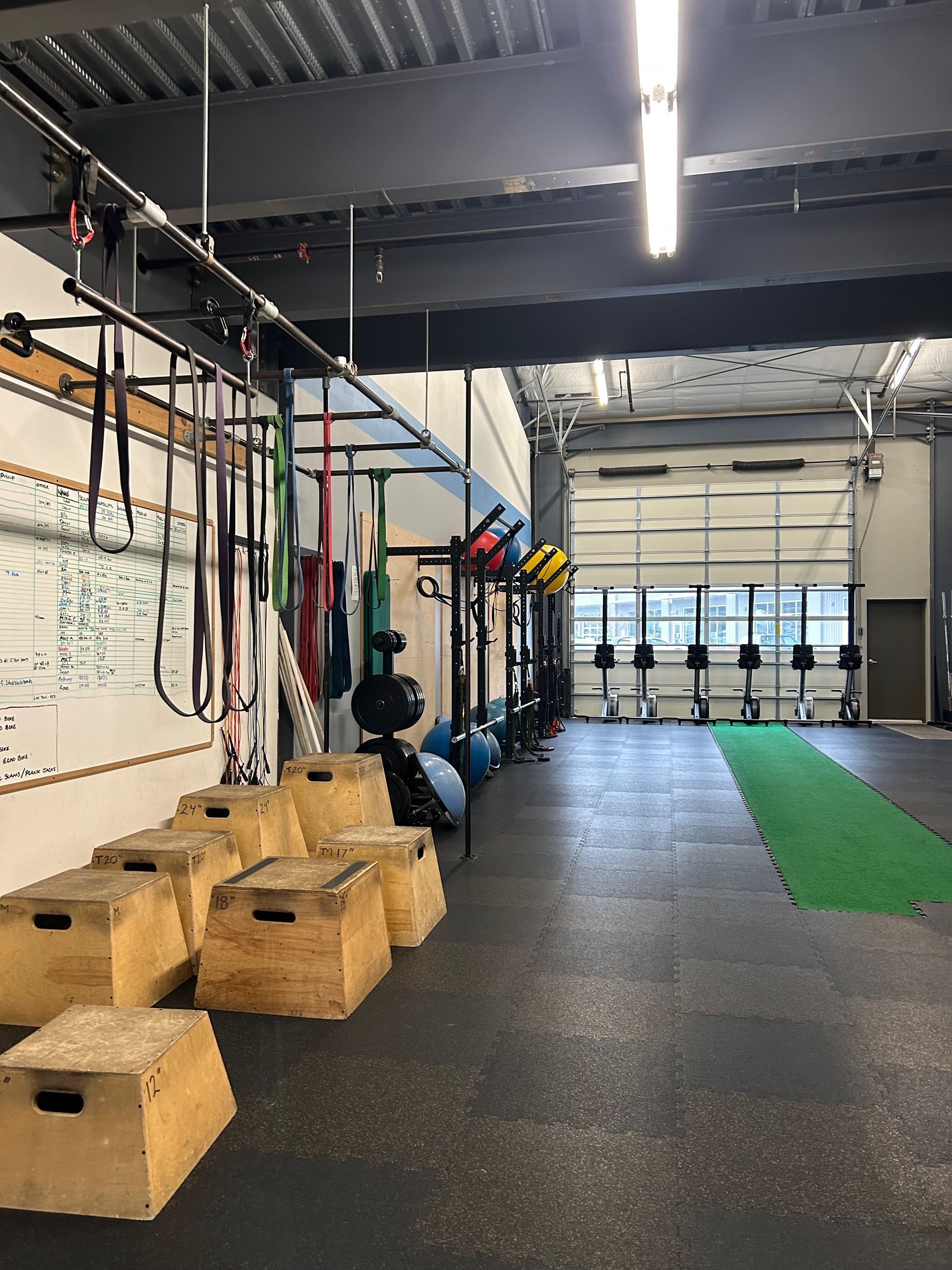 A gym interior with wooden plyo boxes in the foreground, hanging resistance bands, and rowers on a green turf lane.