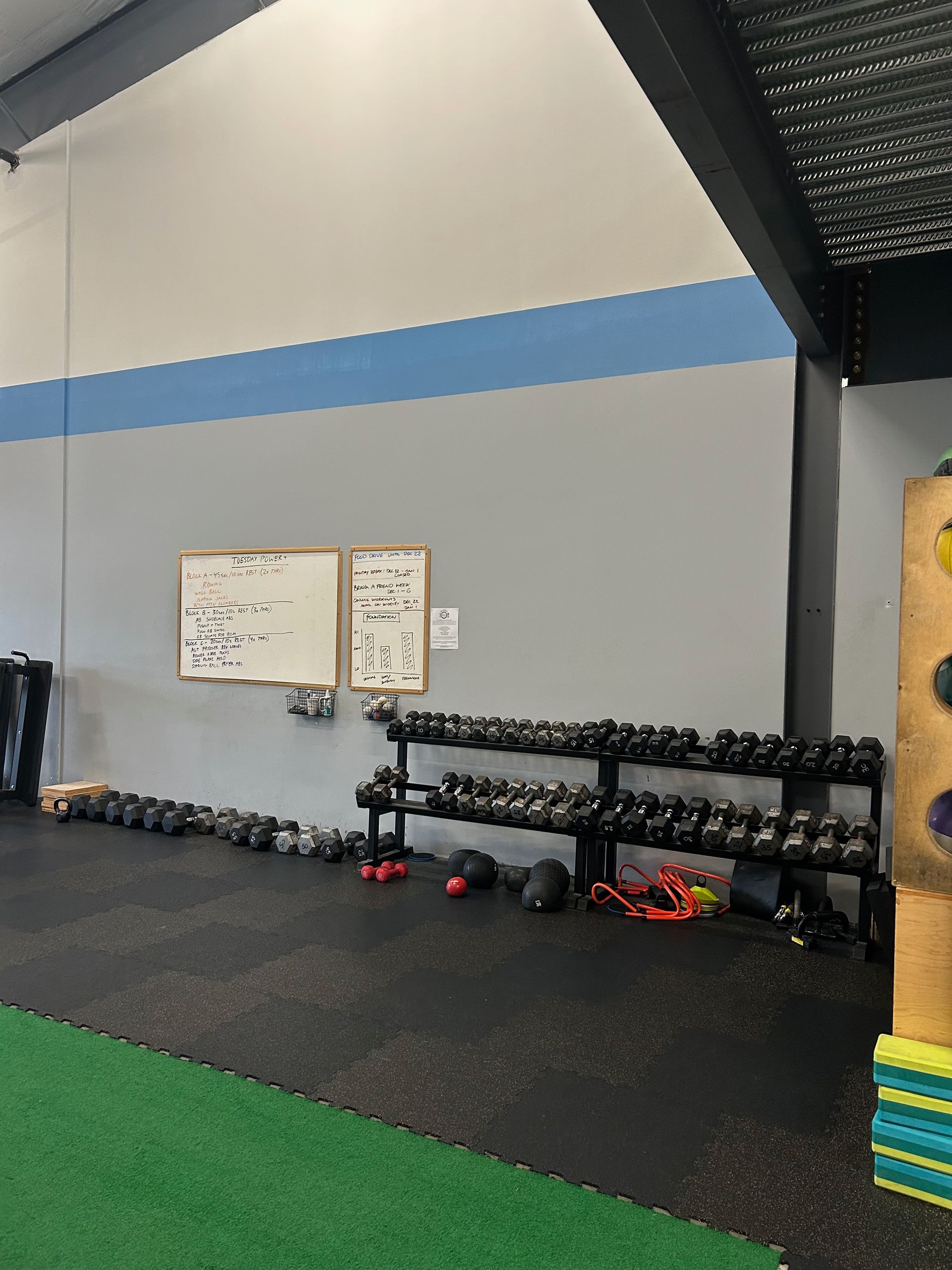 A gym wall featuring a whiteboard, a tiered dumbbell rack, and a row of kettlebells on the floor against a gray wall.