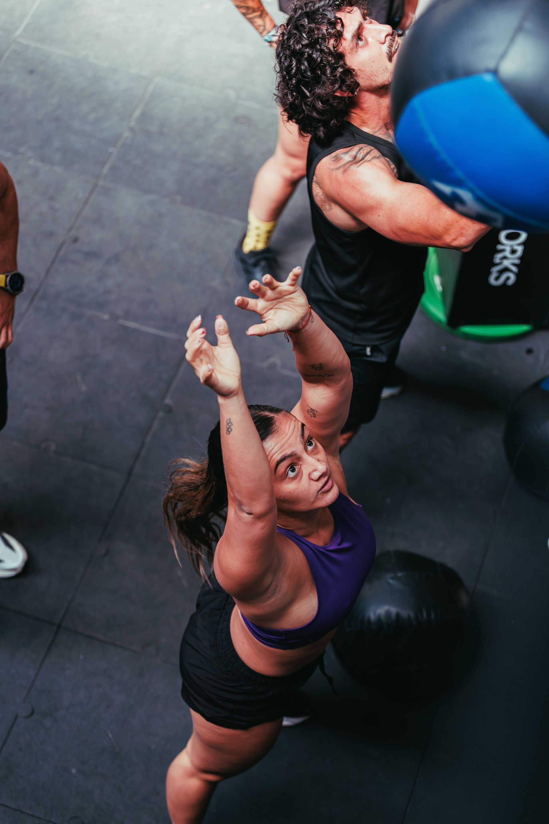 Two people doing medicine ball wall throws in a gym, one in a purple top reaching up, one in black catching.