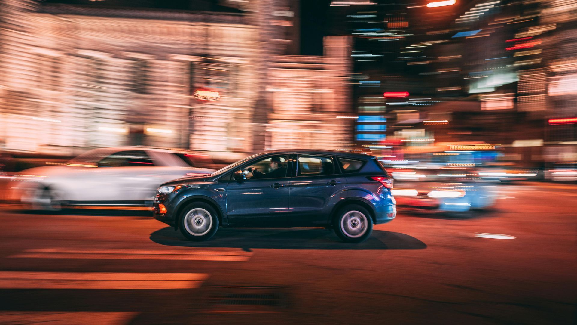Blue car driving on a city street at night with motion blur. Buildings and other cars are visible.
