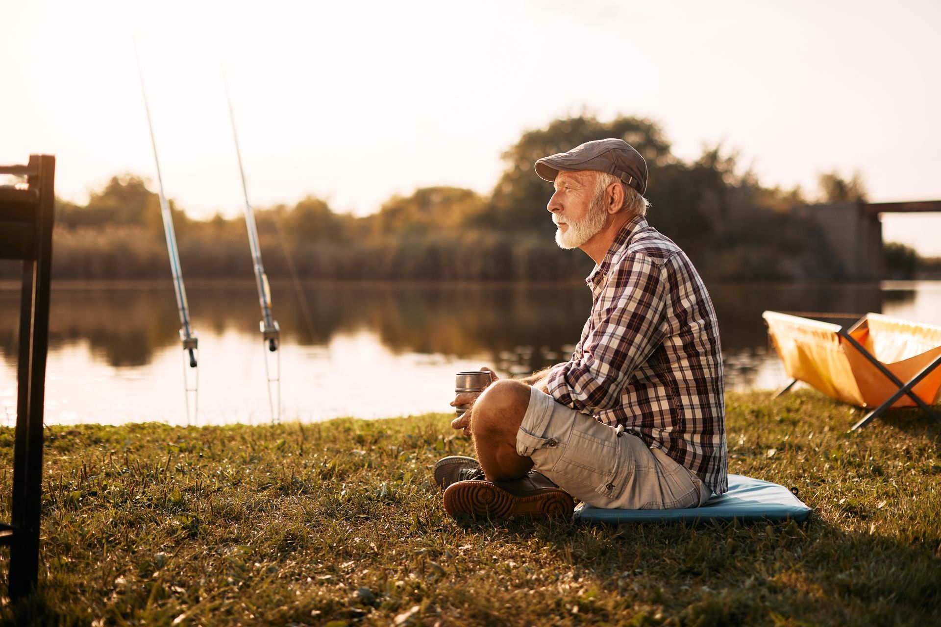 An elderly man is sitting on the grass near a lake with fishing rods.