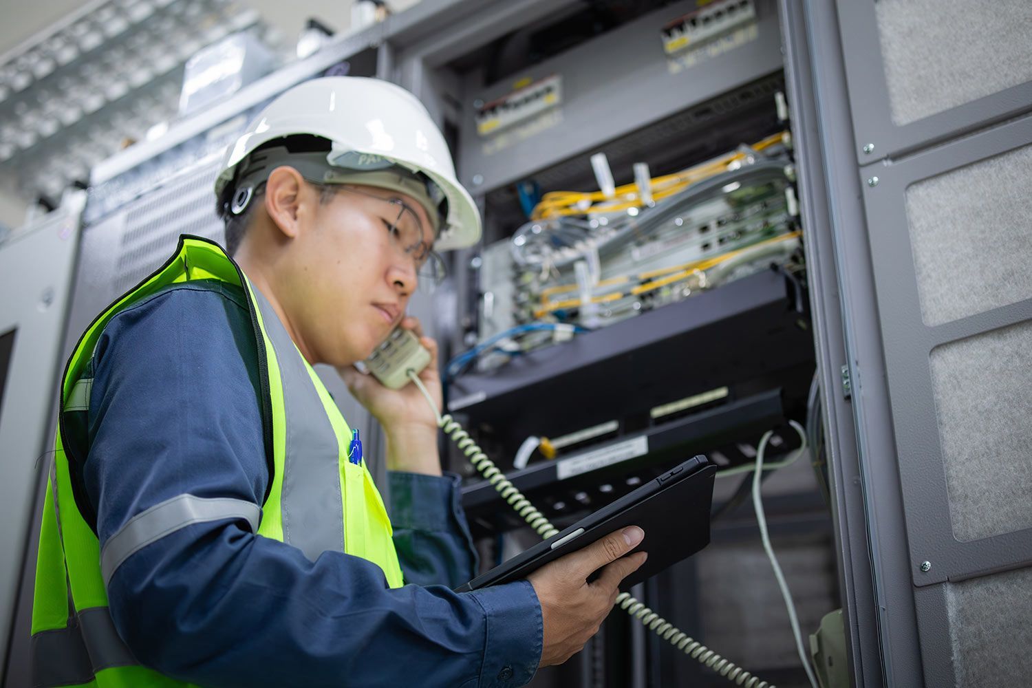 A network technician wearing safety gear holds a tablet and uses a telephone in a server room.