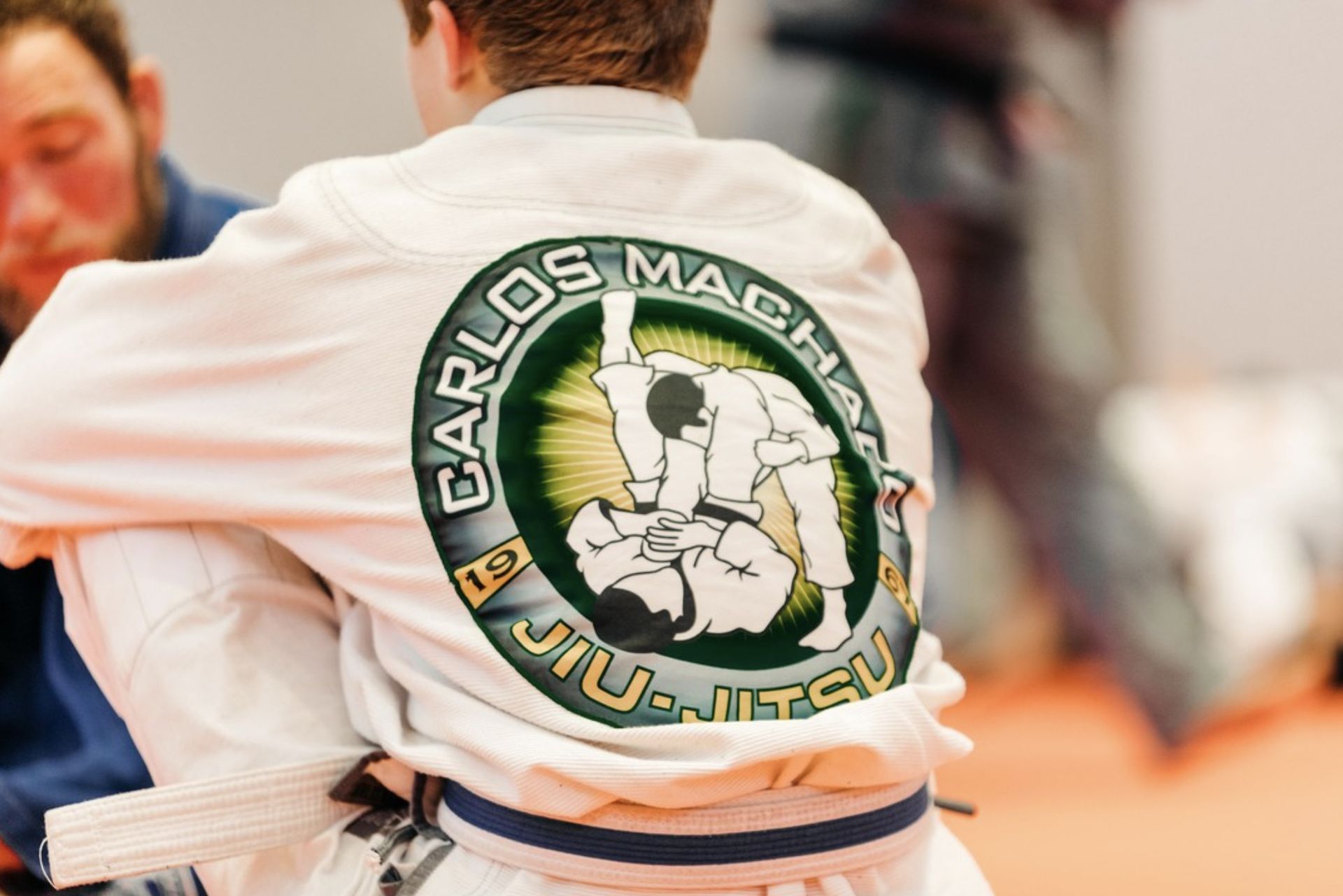 A man sitting with the carlos machado jiu jitsu logo imprinted on his back.