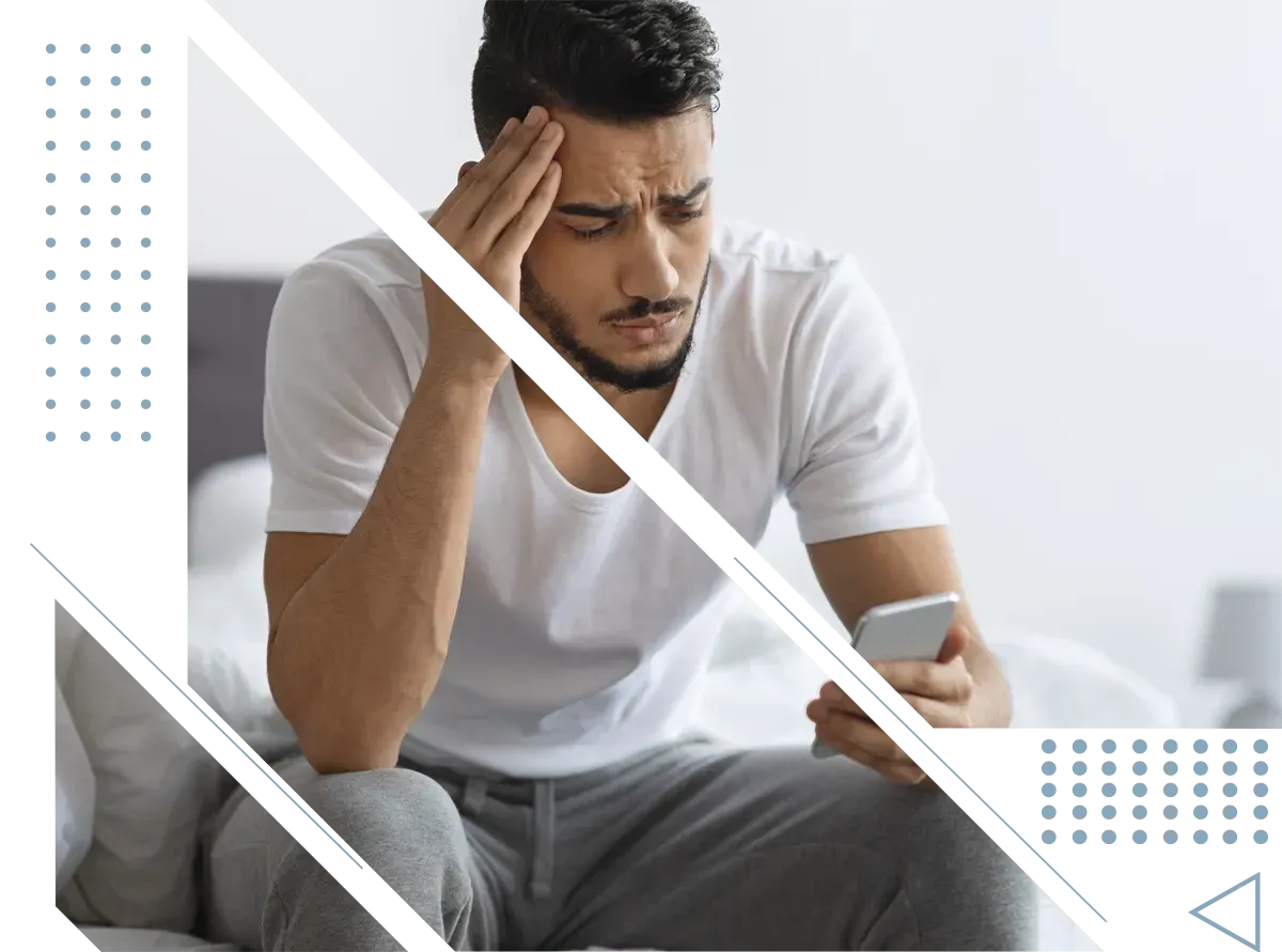 A man is sitting on a bed holding his head while looking at his phone.