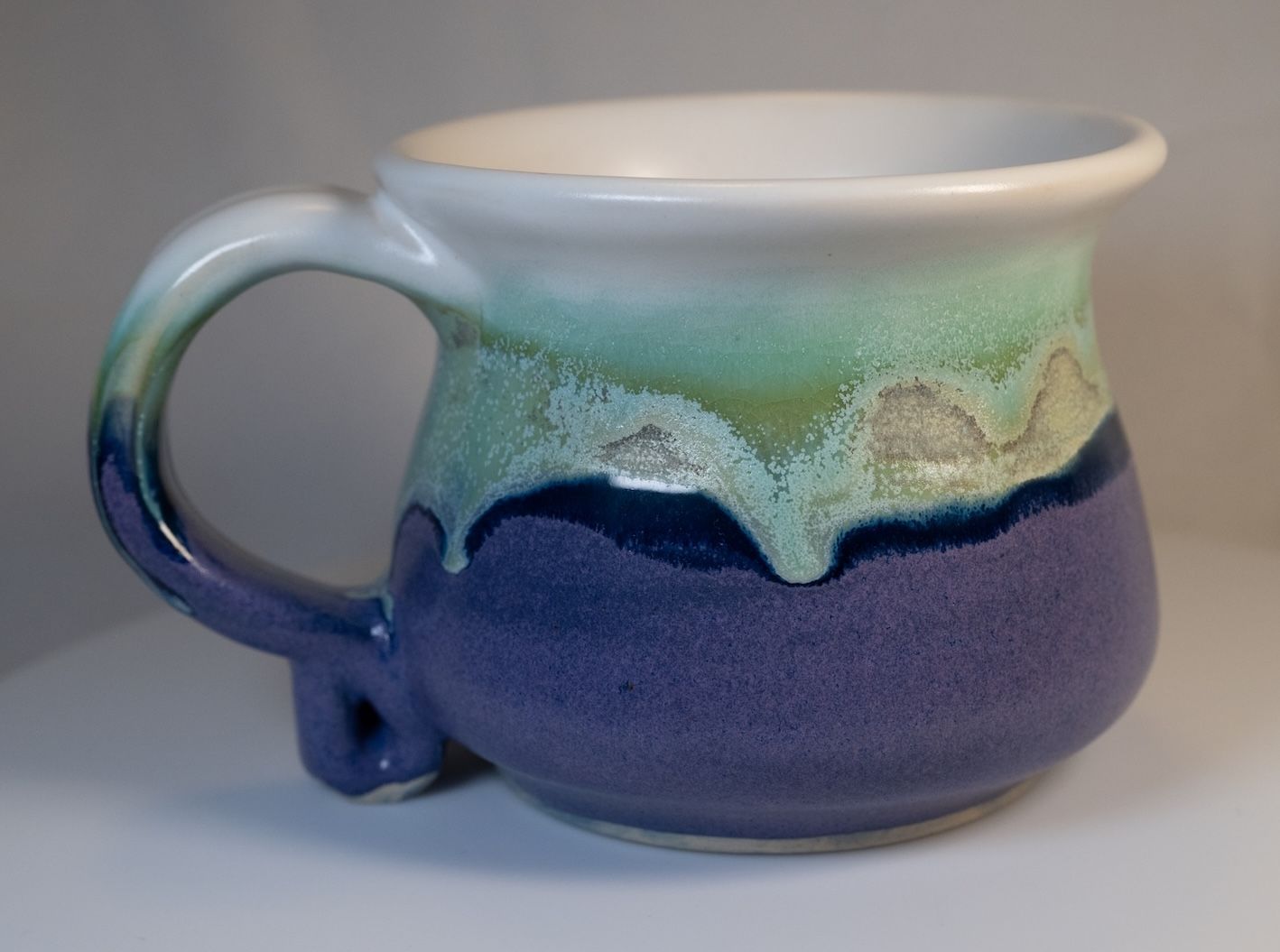 Ceramic mug with layered glaze: white, green, and purple. The handle is blue.