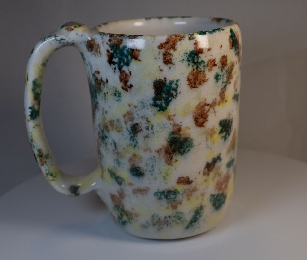 Ceramic mug with speckled brown, green, and yellow patterns on a cream background.