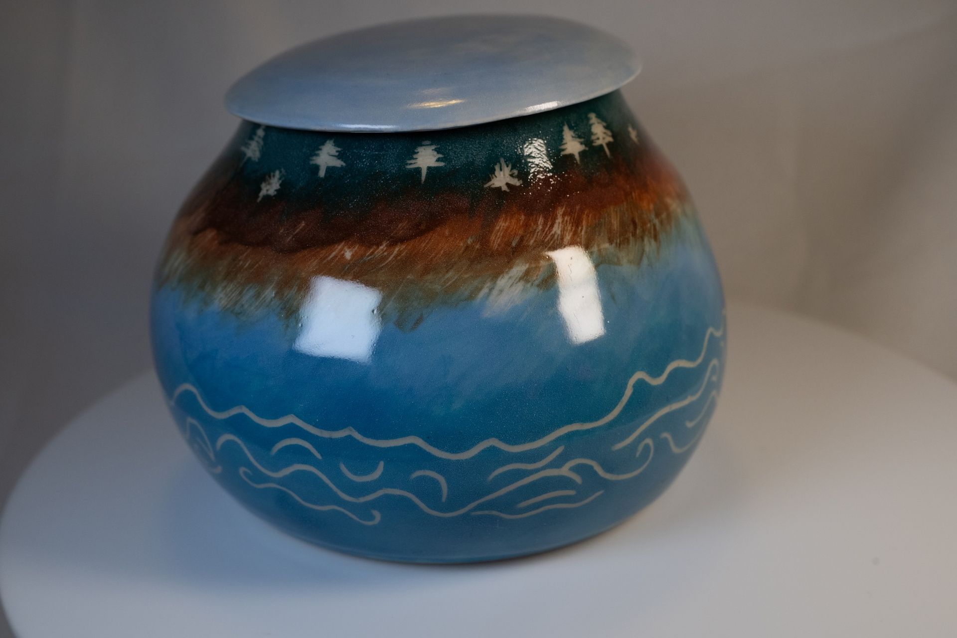 Ceramic urn with blue, brown, and green glaze. Decorative waves, trees, and lid.
