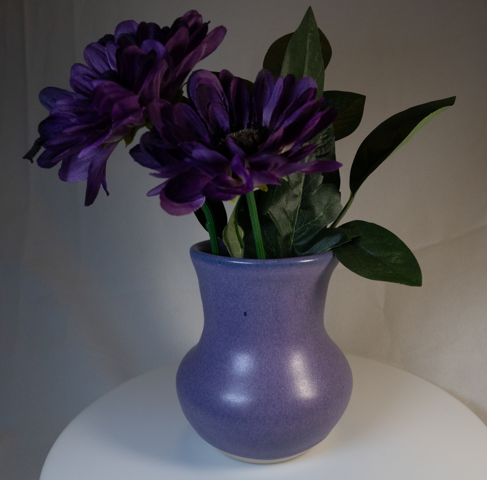 Purple flowers in a lavender vase on a white surface, against a neutral background.