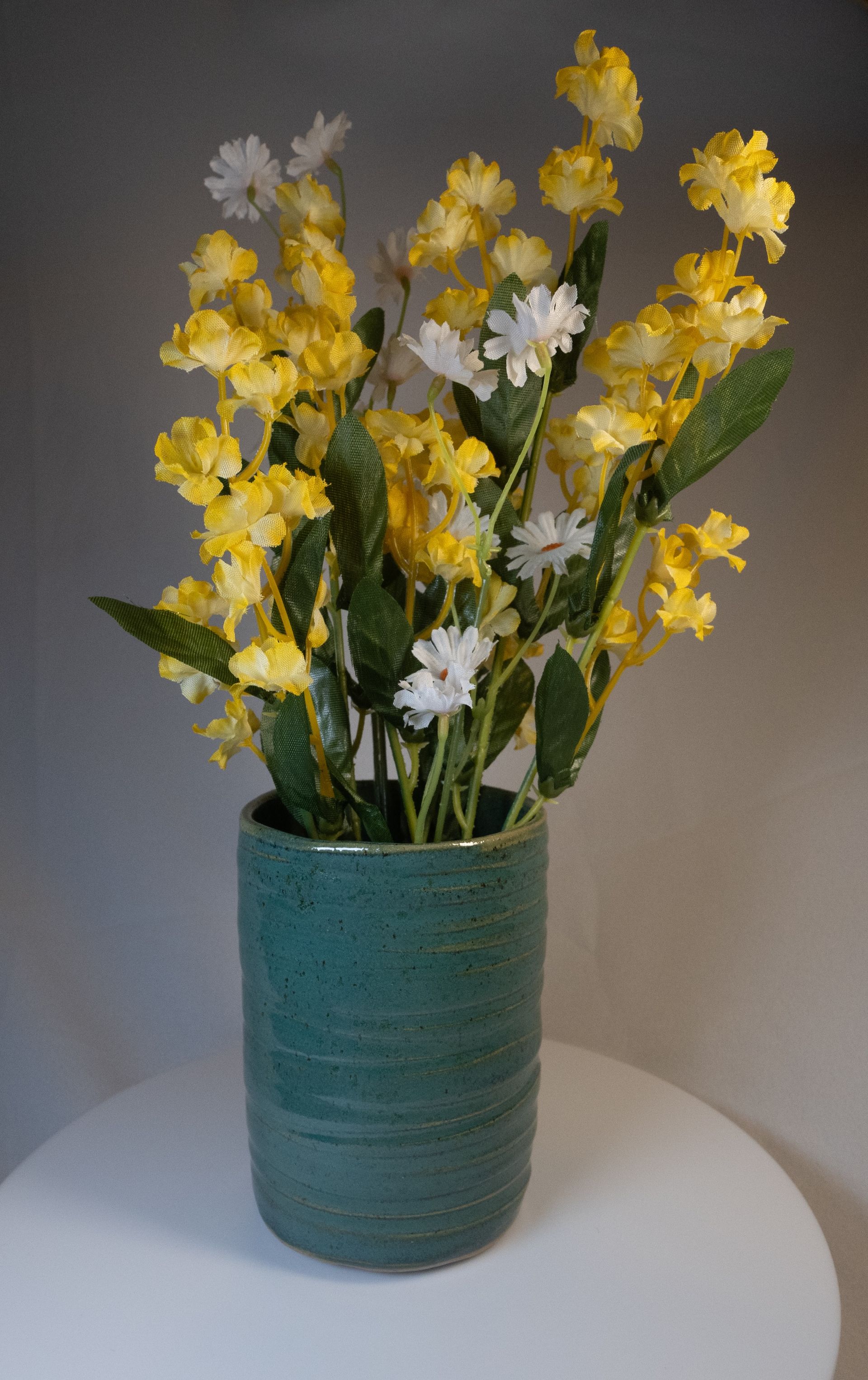 Green vase with yellow and white flowers on a white surface.