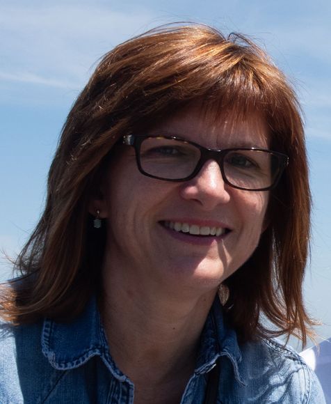 Woman with brown hair and glasses smiling outdoors in a denim shirt.