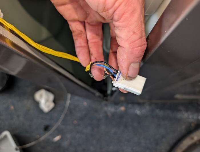 Hand holding a white electrical connector with multiple colored wires near a gray appliance panel.
