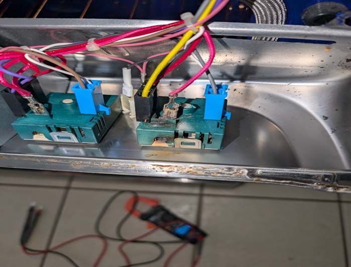 Close-up of appliance control panel with wires, blue connectors, and a multimeter.