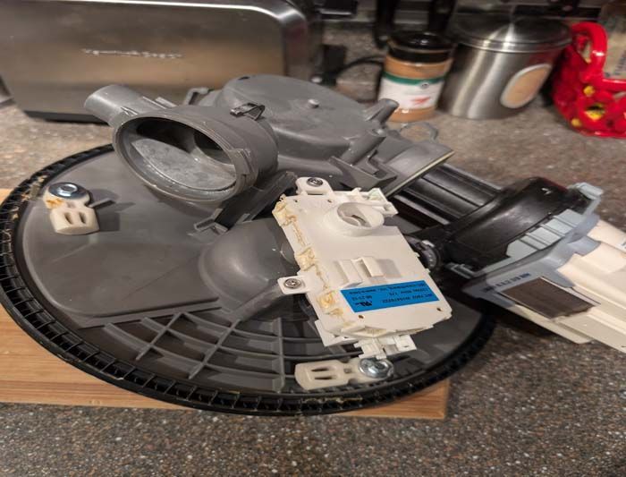 Dishwasher parts disassembled on a countertop. Includes pump, control module, and base with filter.