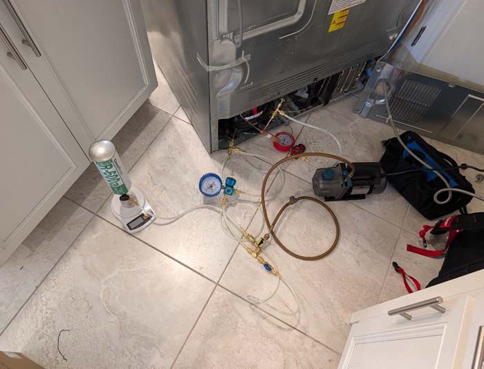 Refrigerator repair: gauges, hoses, vacuum pump, and refrigerant can near the appliance in a kitchen.