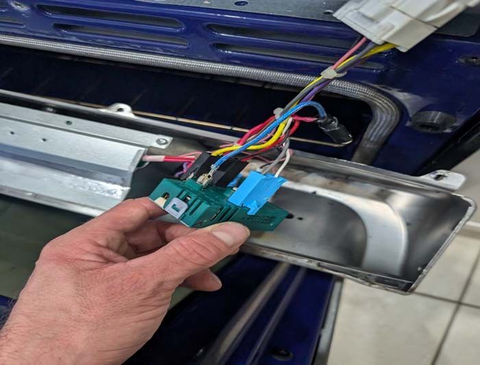 Hand holding an oven component with colored wires connected. It has a green and blue switch. Oven interior.