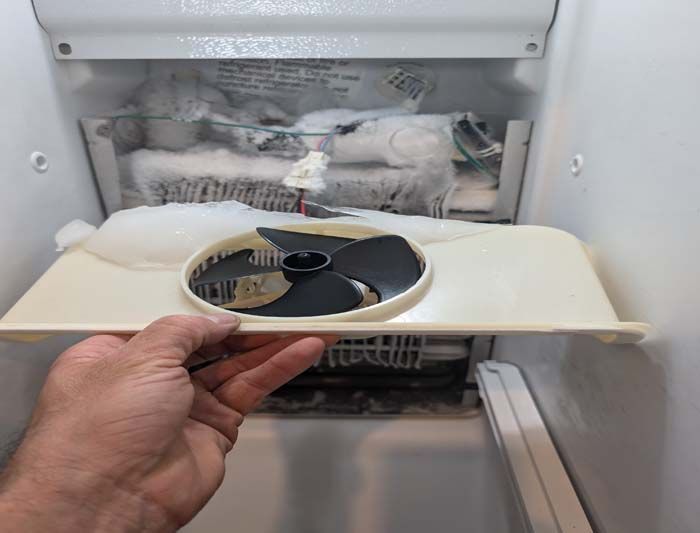 A hand holding a shelf with a black fan, inside a heavily frosted refrigerator freezer.