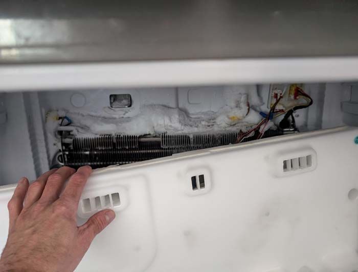 Hand holding a white panel inside a freezer, exposing a frozen coil. Wires and vents are visible.