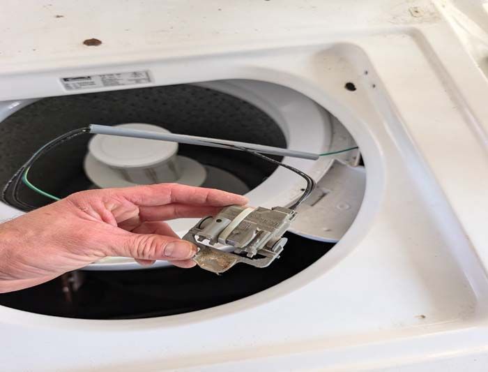 Hand holding a washing machine lid lock switch, with the machine's open interior in the background.