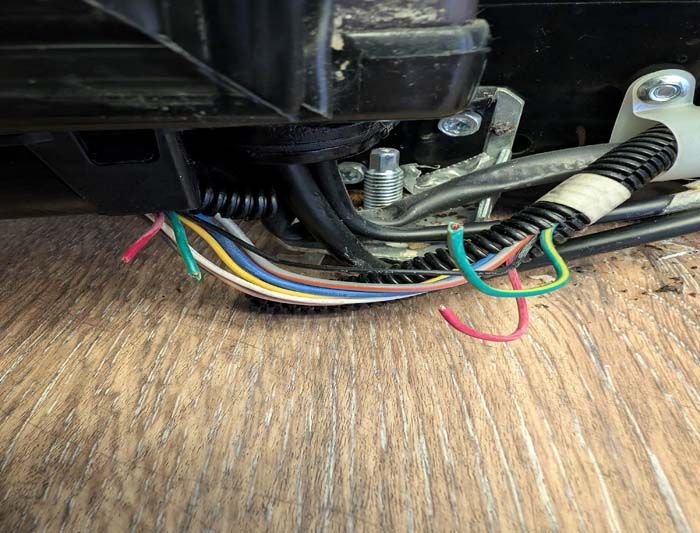 Close-up of exposed multi-colored wires beneath a black component, likely from a machine or appliance, on a wooden surface.