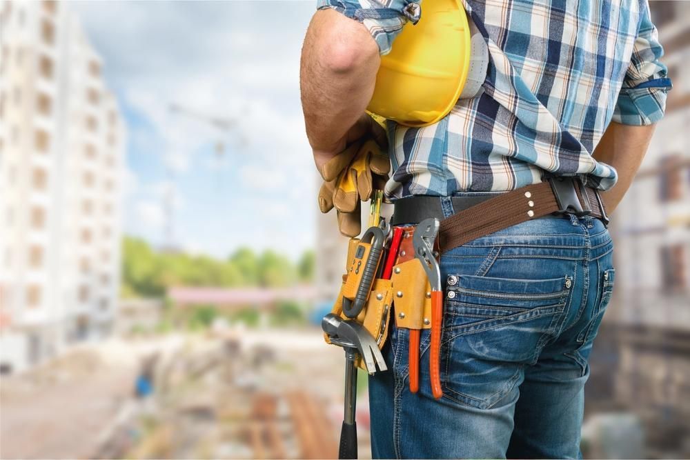 Construction Worker In Blue Jeans And Plaid Shirt, Holding Yellow Helmet And Tools At A Construction